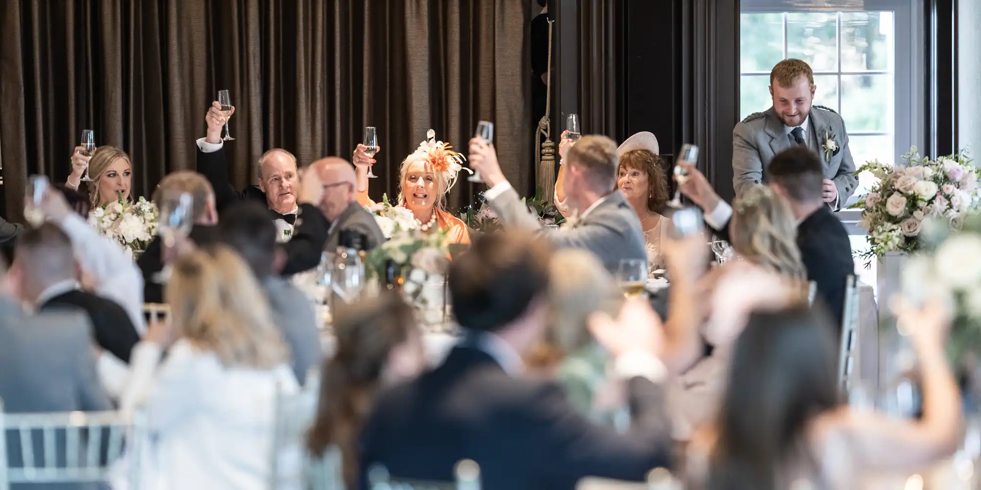 A group of people seated at tables raising their glasses in a toast during an indoor event. The background features a curtain and floral arrangements.