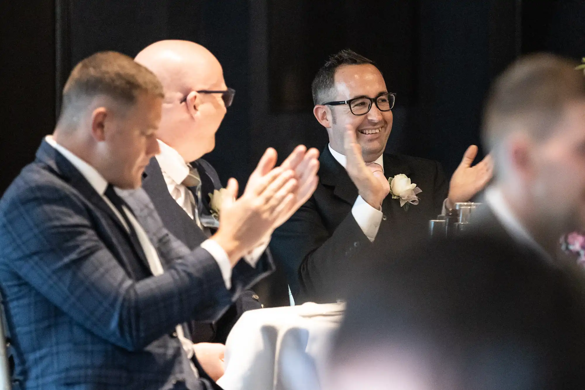 Three men in suits are seated at a table, clapping and smiling. They appear to be at a formal event or celebration.