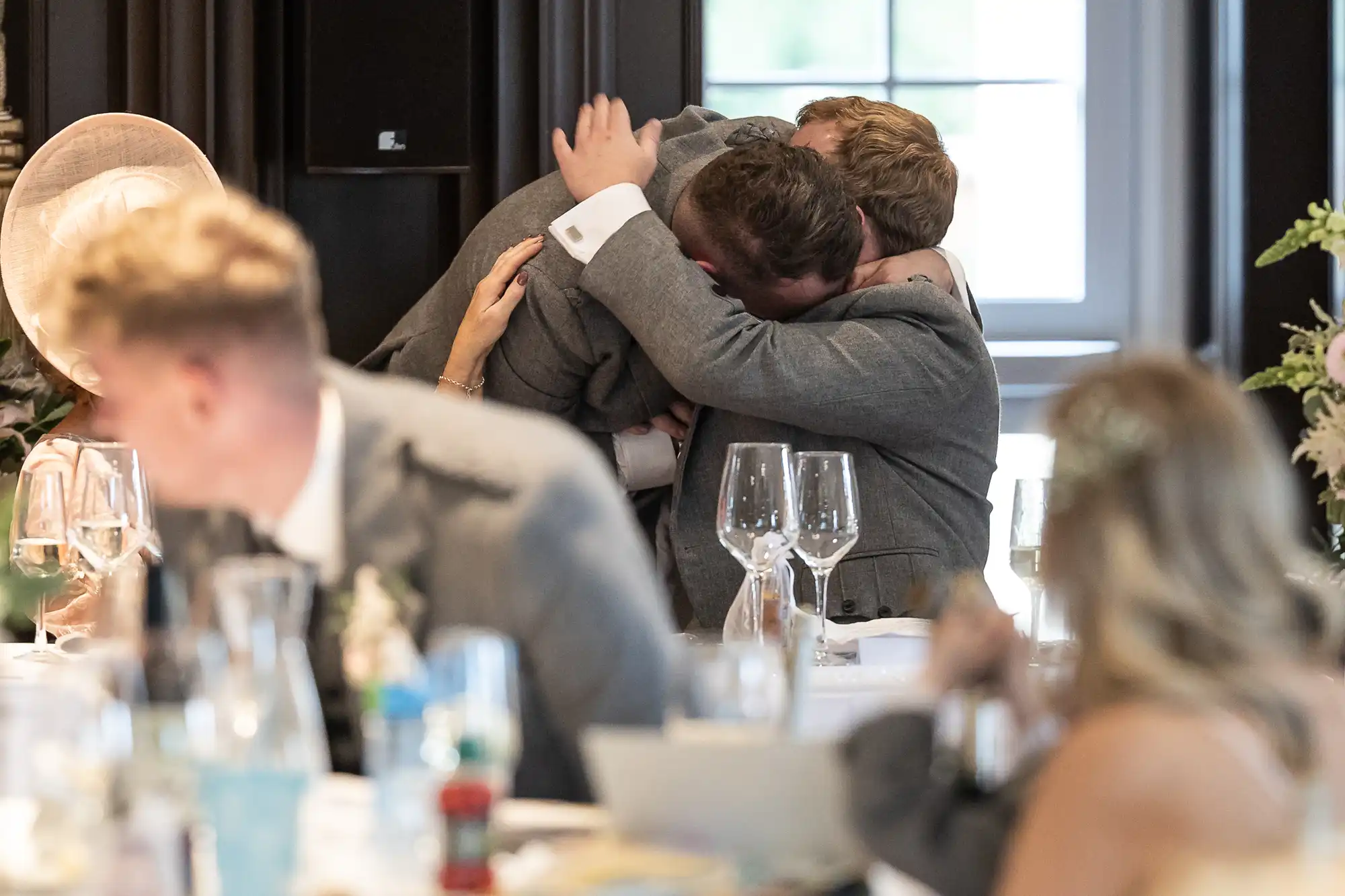 Two people in grey suits embrace emotionally at a formal event, while seated guests in the foreground look on.