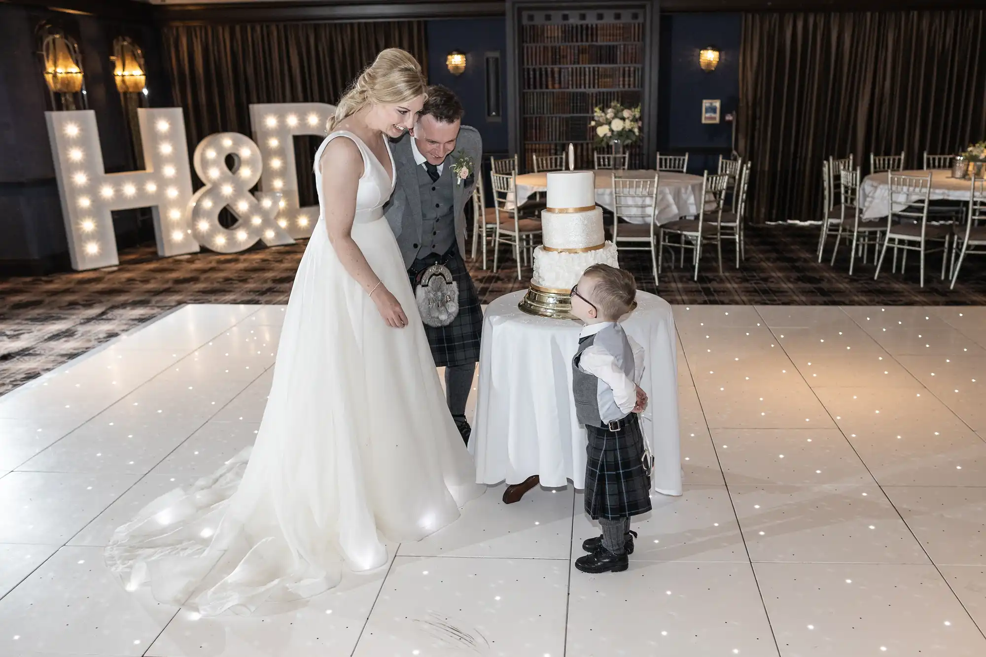 A bride and groom assist a young boy as he looks at a three-tiered wedding cake on a stand, with large illuminated letters "H & D" in the background.