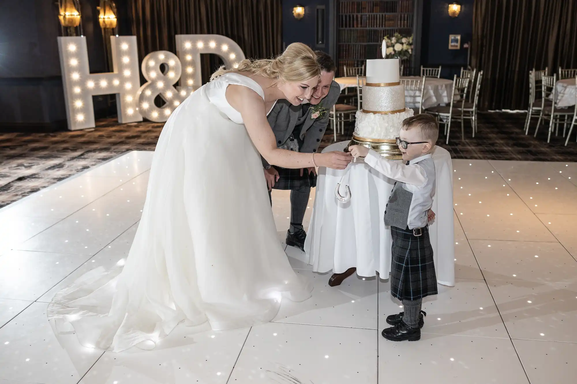 A bride in a white gown and a groom in a kilt help a young boy in a kilt cut a three-tiered wedding cake at a wedding reception. Large lit initials "H & D" are visible in the background.