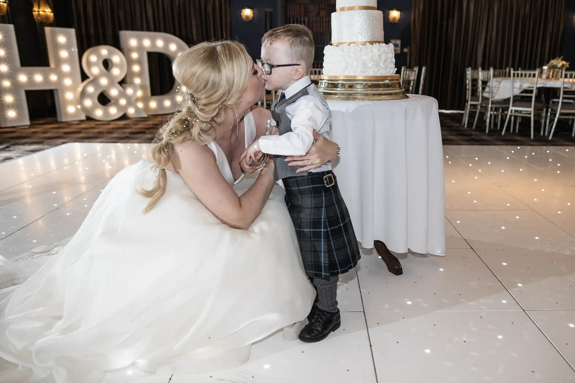 A woman in a white dress kisses a young boy in a kilt on the dance floor near a table with a multi-tiered cake. Large illuminated letters "H&D" can be seen in the background.