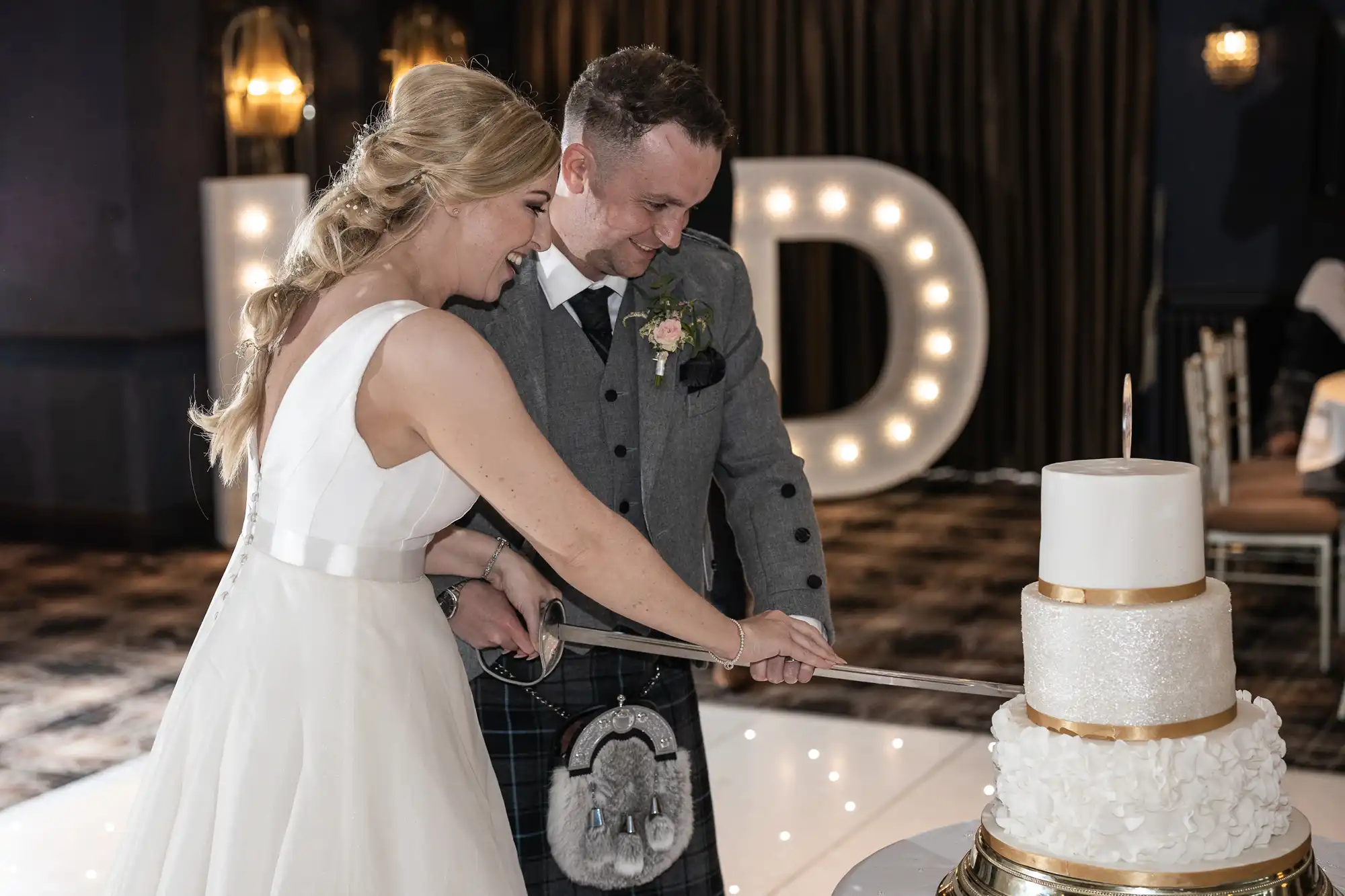 A bride in a white dress and a groom in a gray suit cut a tiered wedding cake together with a sword. The background features large illuminated letters.