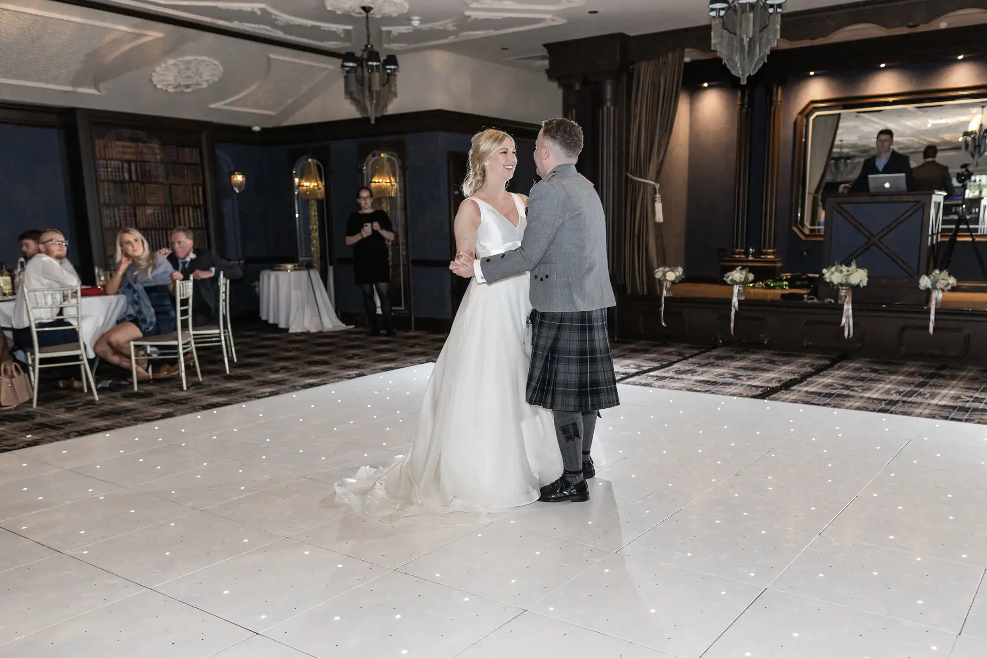 A bride in a white dress and a groom in a kilt have their first dance at a reception, with a few guests seated at tables in the background and a DJ in a booth on the stage.