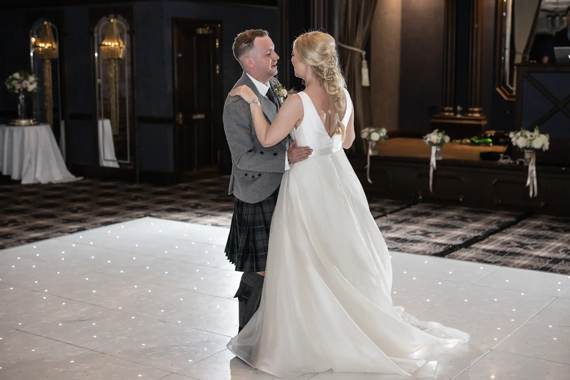 A couple shares their first dance at a wedding reception. The man wears a kilt, and the woman is in a white wedding gown. They are on a lit dance floor with a dimly lit background.