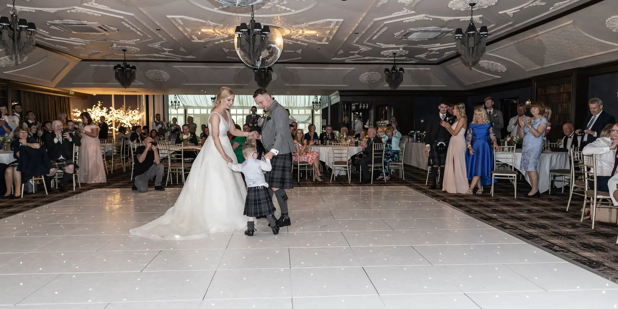 A bride, groom, and small child in kilts dance together in the center of a room, while guests seated at tables watch and take photos.