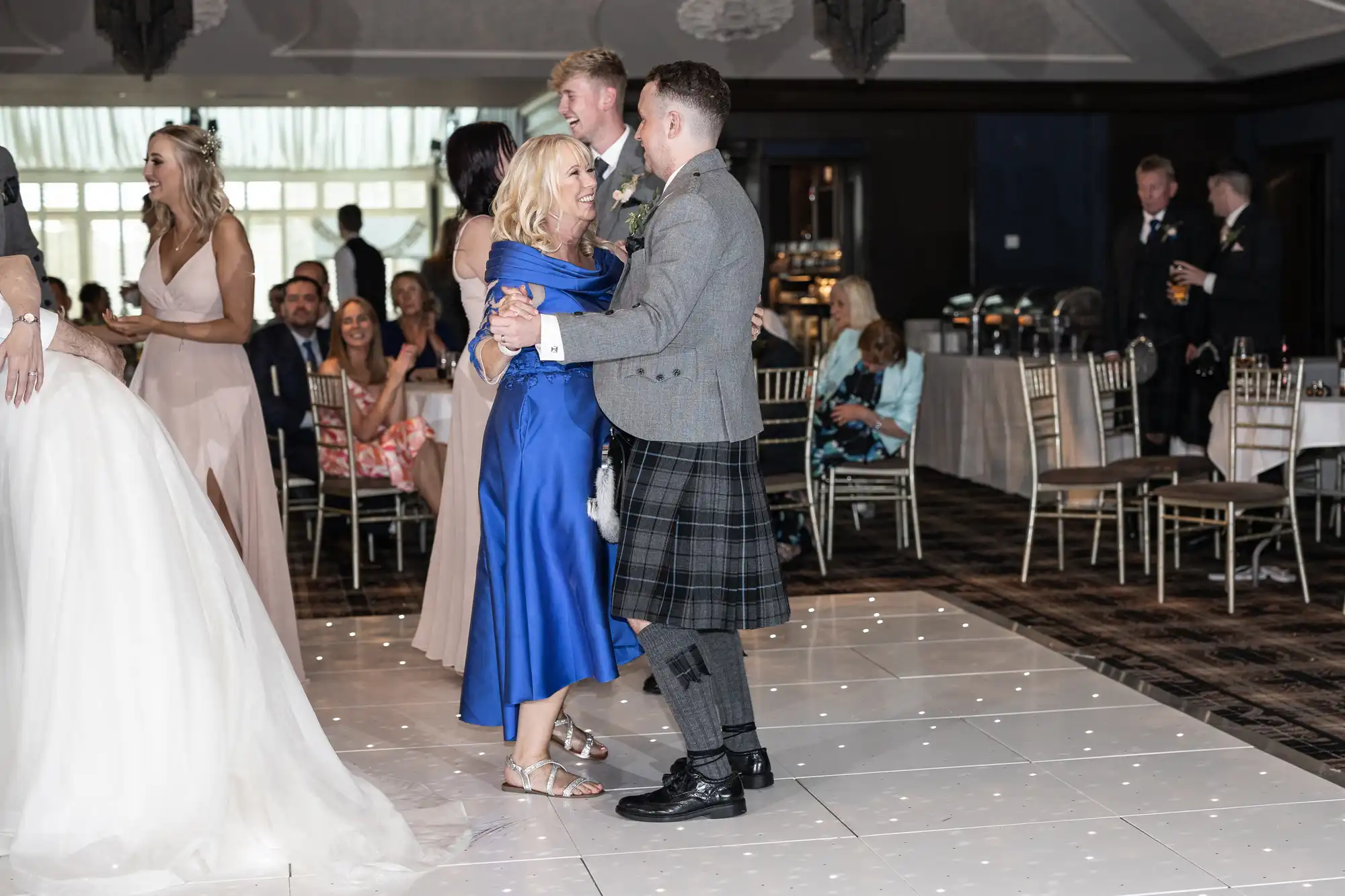 A couple, dressed in formal attire, dances on a white-lit dance floor at a wedding reception while others sit and stand in the background.