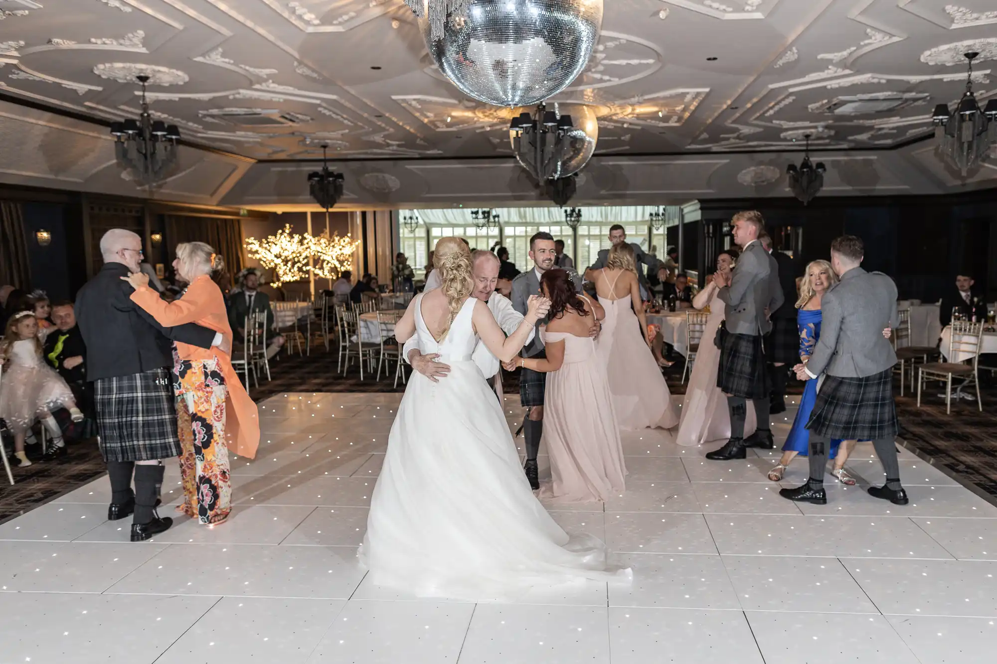 A wedding reception with couples dancing on a white-lit dance floor under a large disco ball in an elegantly decorated venue. Guests are dressed formally, with some men in kilts.