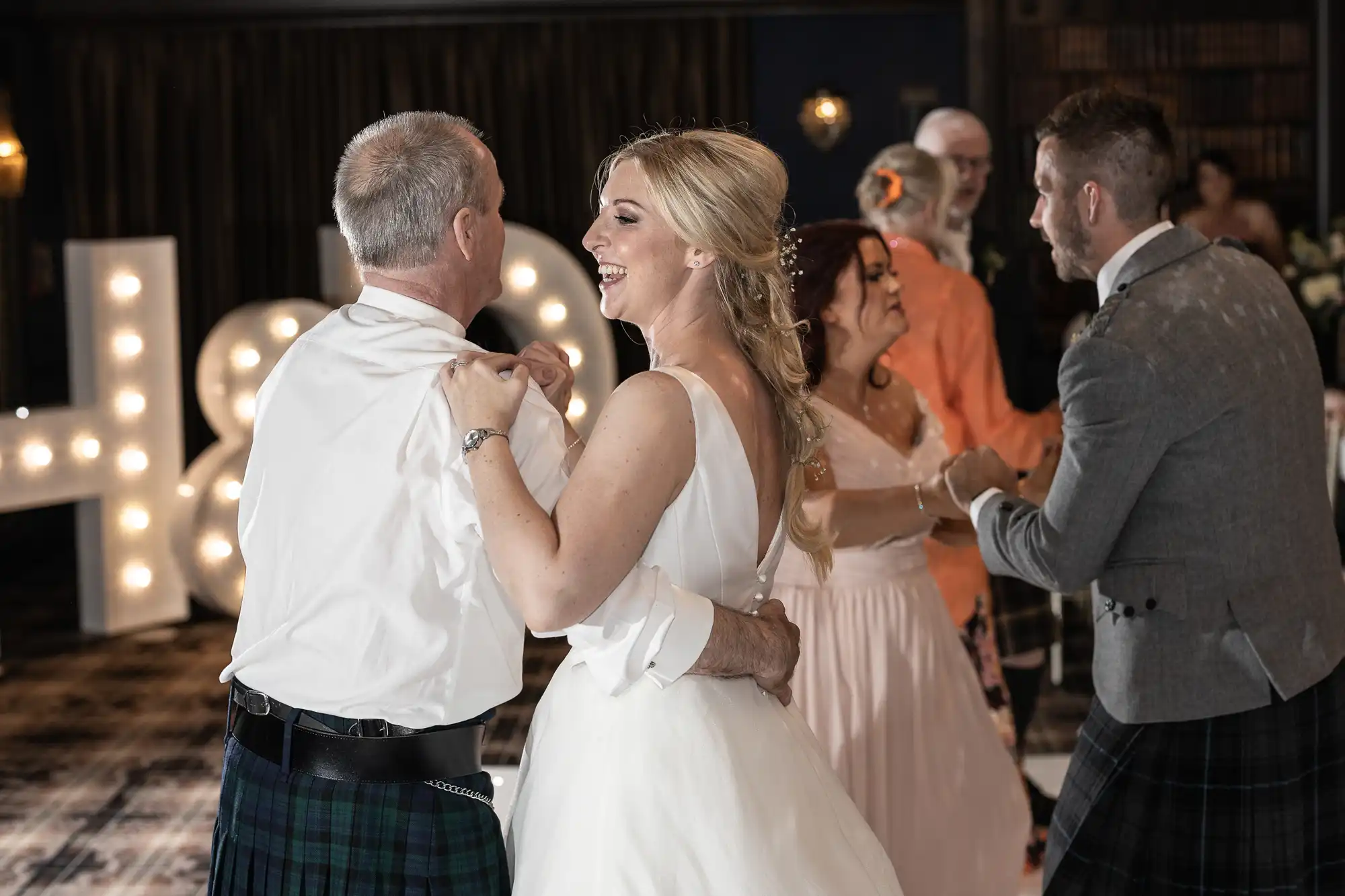 A bride and an older man dance together in a festive room, surrounded by other dancing couples. She is smiling and they are both wearing formal attire.