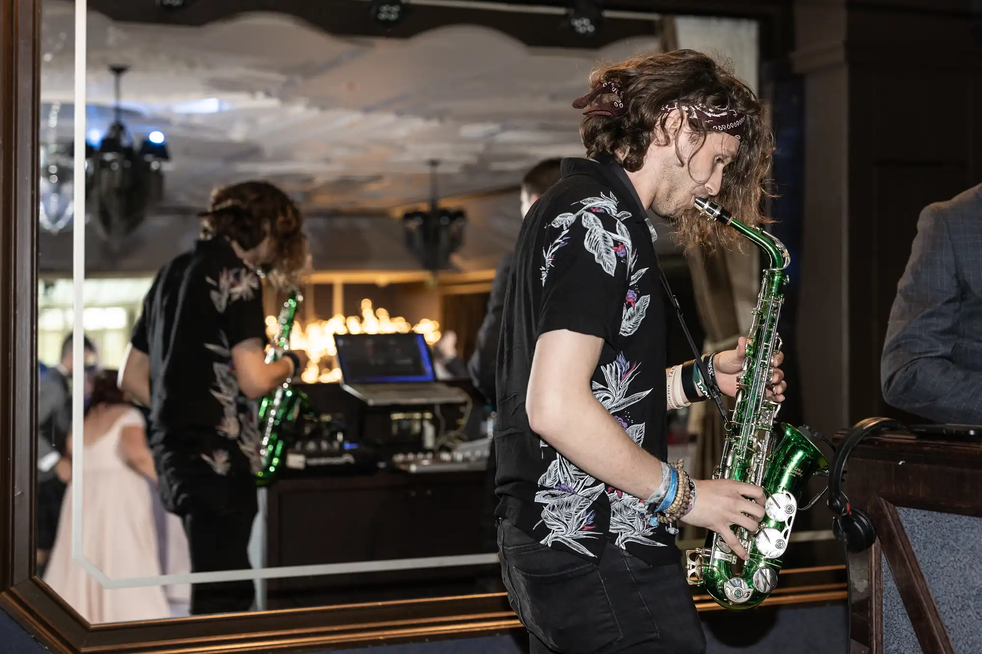 A man with long hair wearing a black floral shirt plays a green saxophone in front of a mirror, with a DJ setup and reflection visible in the background.