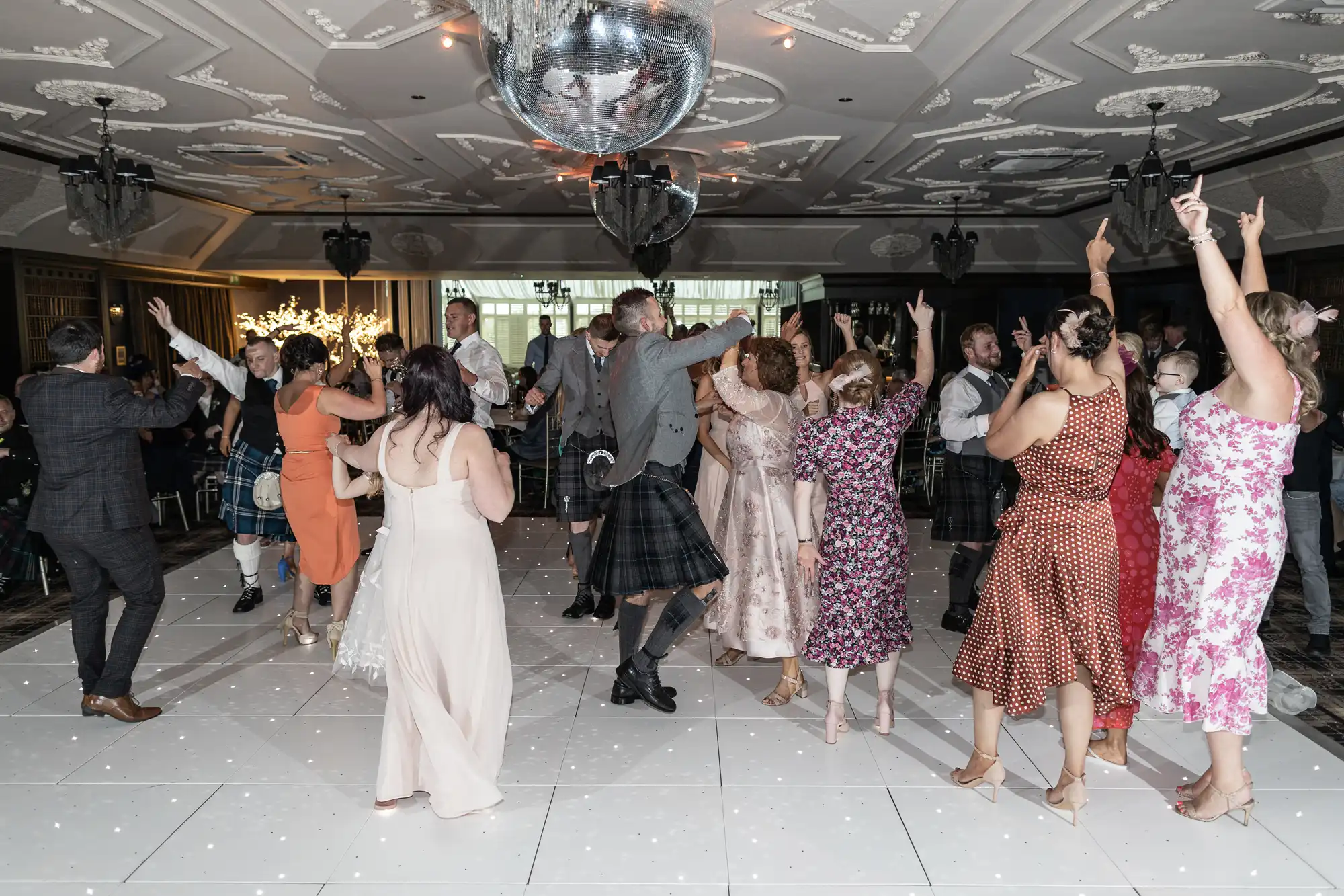 A group of people dance under a disco ball in a formal event hall, dressed in various formal and semi-formal attire, including some in kilts and dresses. The floor is white, and the ceiling is ornate.