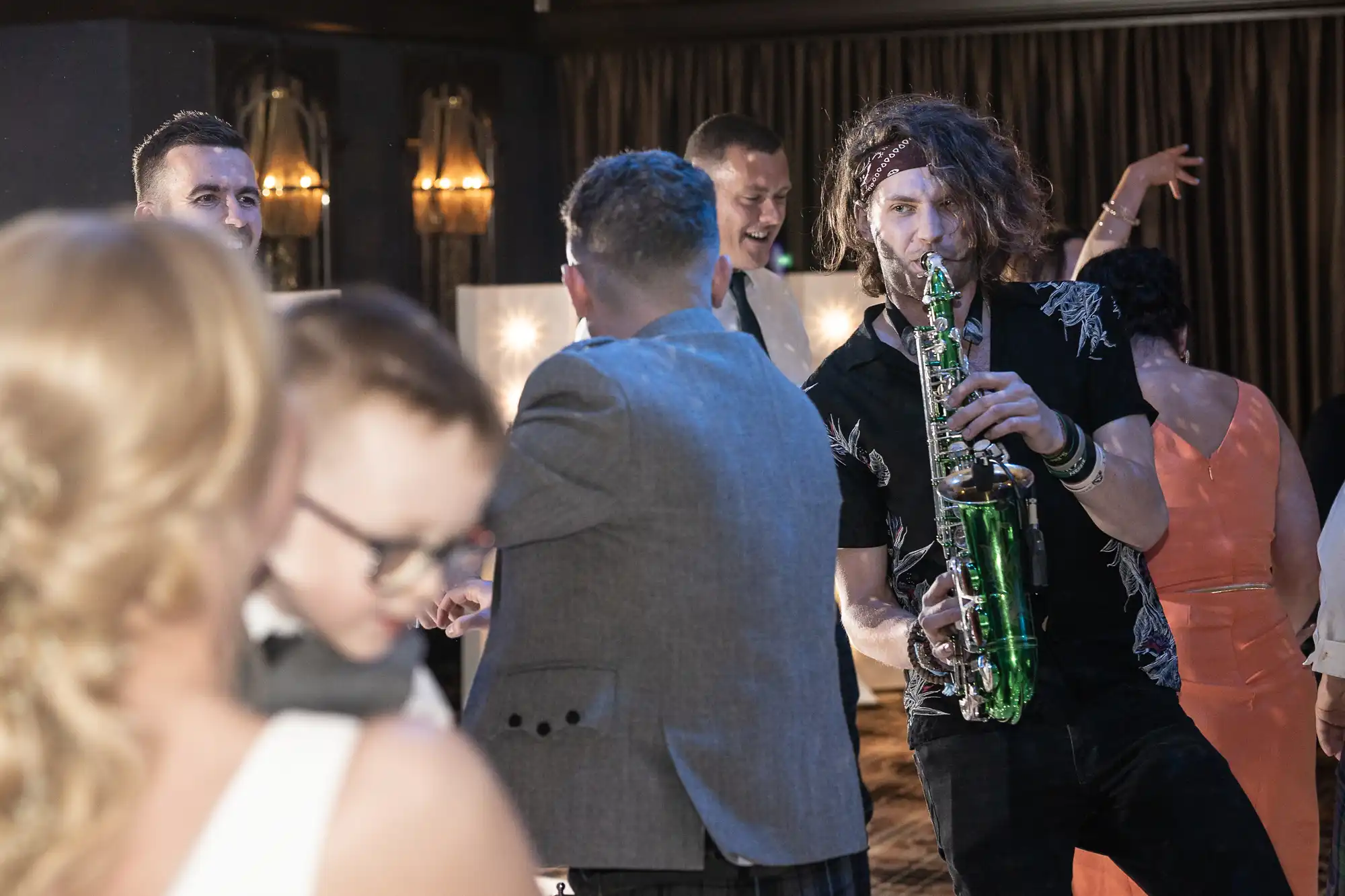 A man with curly hair plays a green saxophone at a lively indoor event. Several other people are engaged in conversation and dancing in the background.