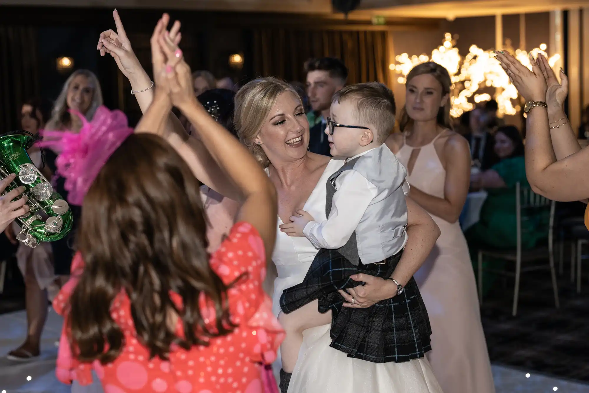 A woman in a white dress holds a child in a kilt while dancing with others at an indoor event.