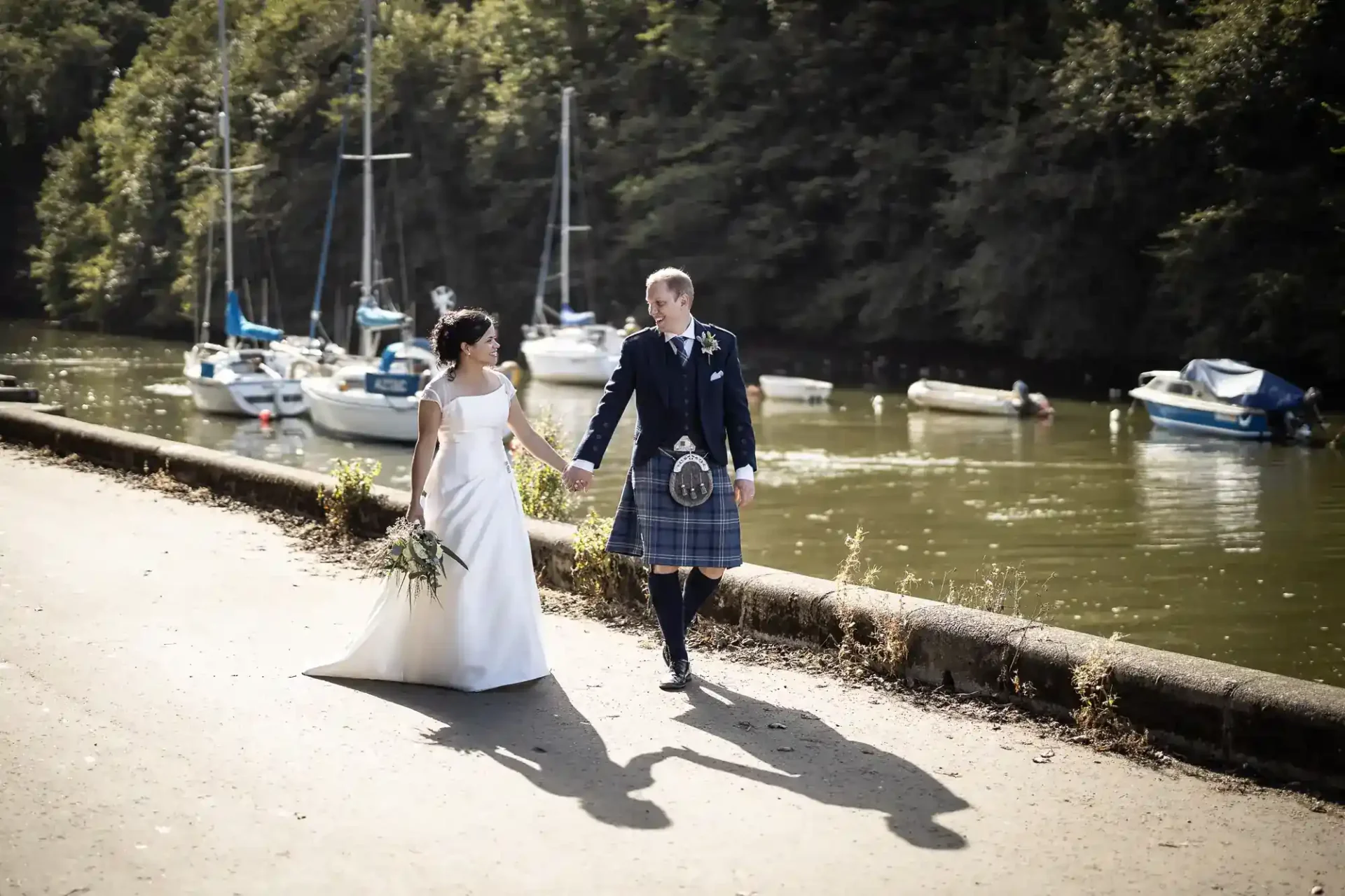 A bride and groom walk hand in hand beside a river with docked boats, the groom in a kilt and the bride holding a bouquet, with trees in the background.