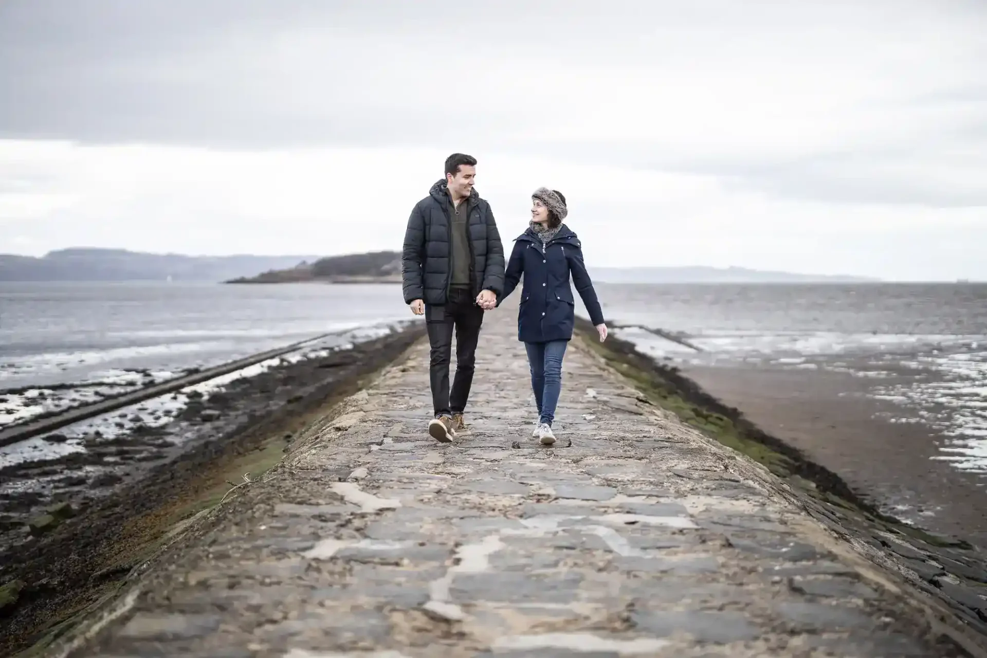 A man and a woman walk hand in hand along a stone path by the sea on a cloudy day.