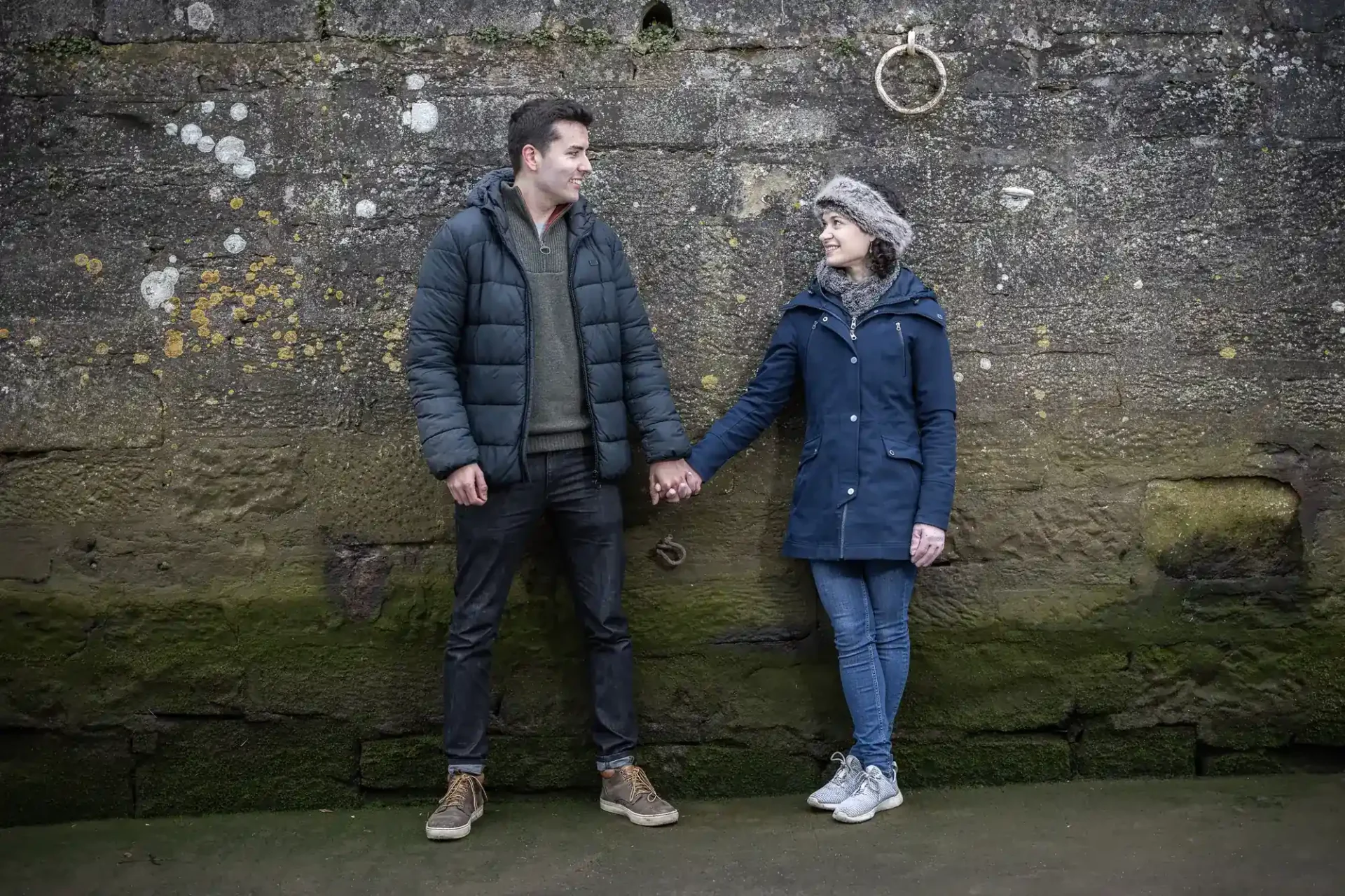 A man and a woman stand against a mossy stone wall, holding hands and smiling at each other. Both wear winter jackets, jeans, and casual shoes.