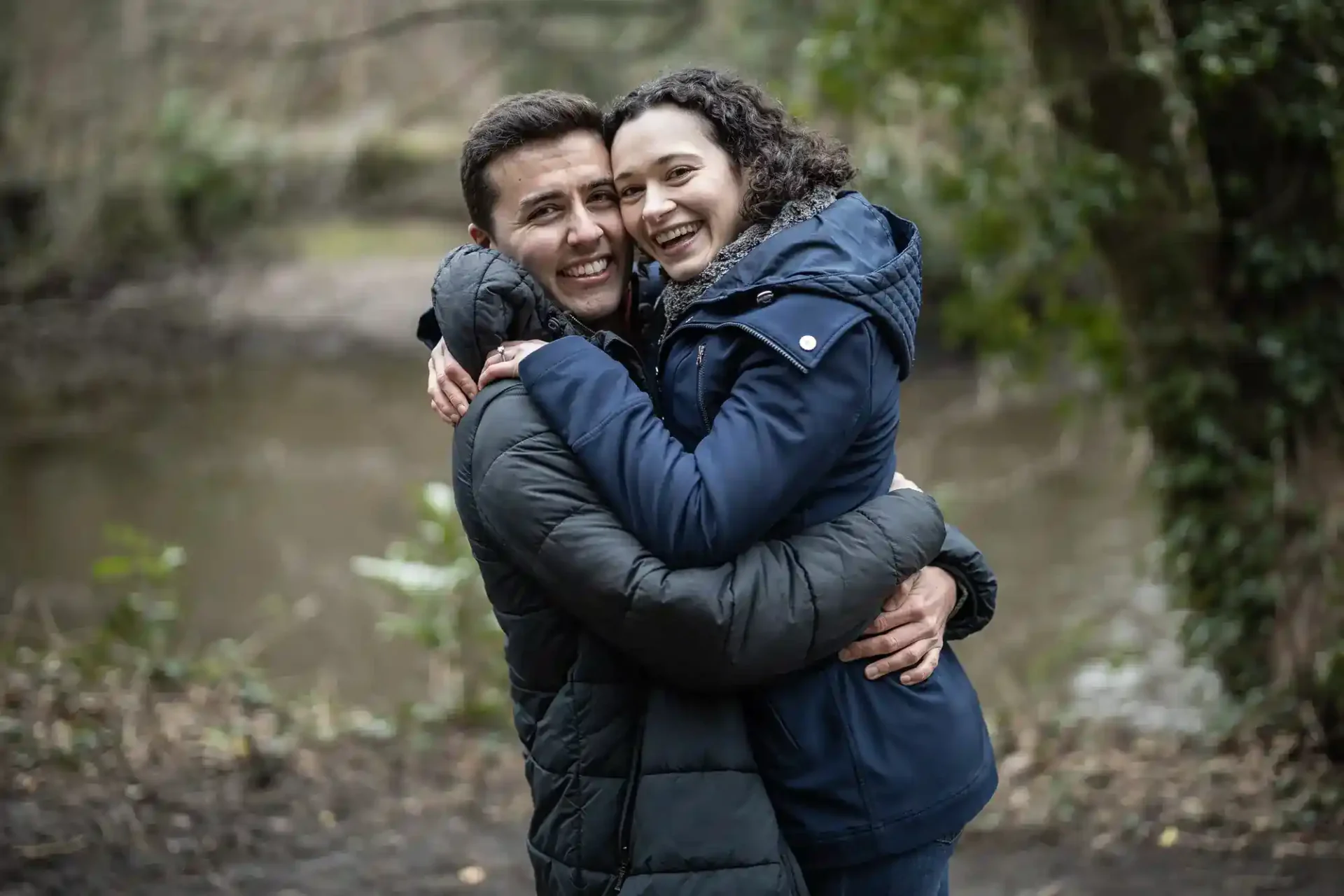 A man and woman in jackets embrace and smile at the camera while standing outdoors near a river with trees in the background.