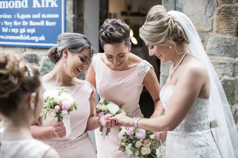 Three women in pastel dresses holding flower bouquets and smiling together. The woman on the right is wearing a wedding dress and veil. They are standing near a stone wall with a blue sign in the background.