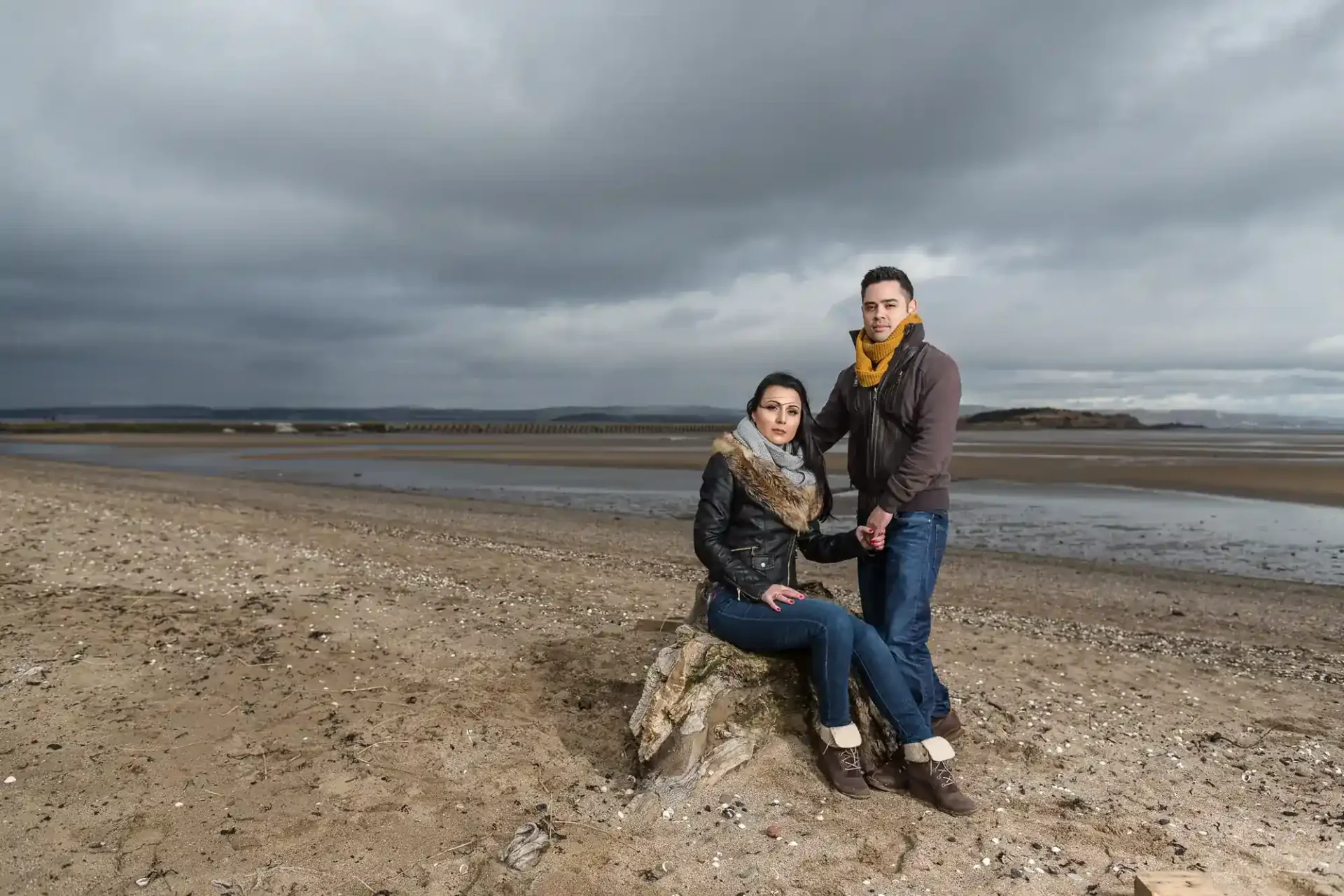 A man and a woman pose together on a cloudy beach; the woman is seated on a rock while the man stands beside her, holding her hand.
