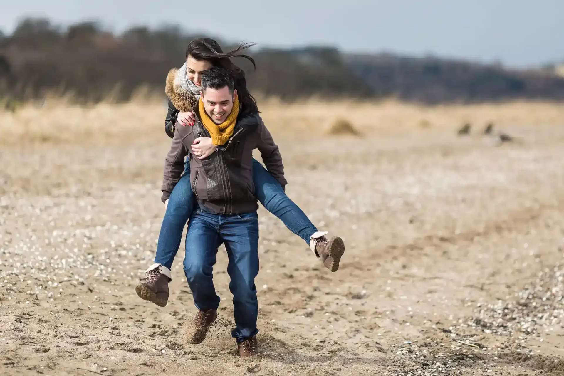 A man gives a woman a piggyback ride on a sandy beach, both are smiling and dressed in casual winter clothing.