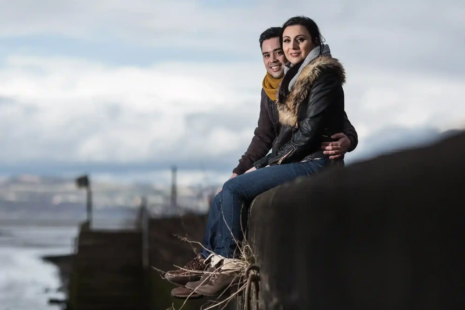 Two people sit on a stone ledge outdoors, with a cloudy sky and distant landscape in the background. The man has his arm around the woman.