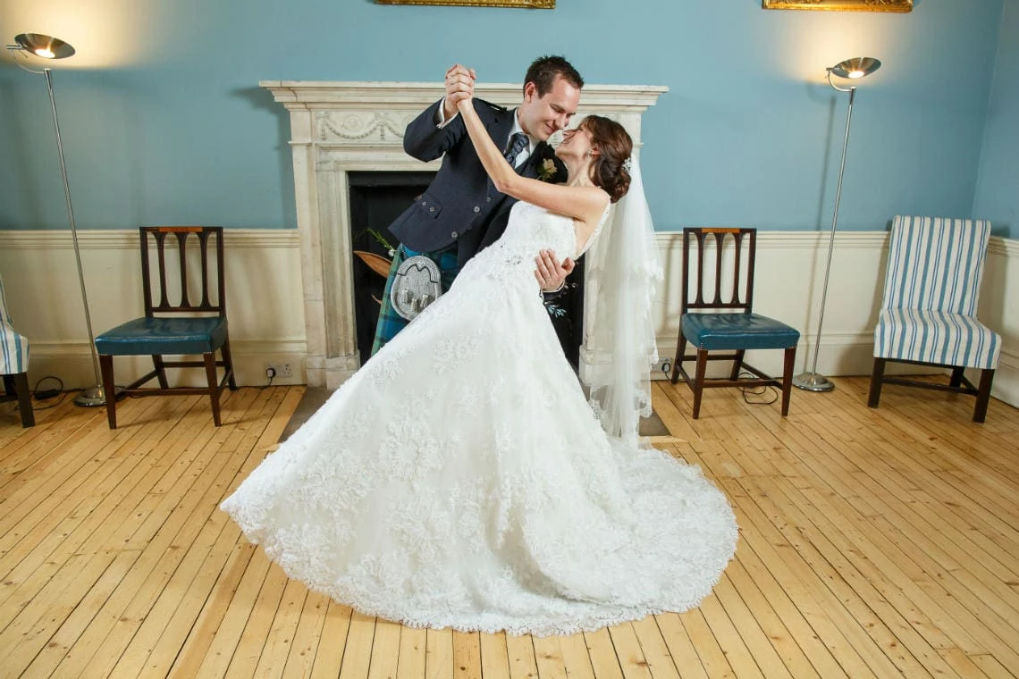 A bride and groom in a dance pose inside a room with a fireplace and striped chairs, the bride in a long white dress and the groom in a navy suit.