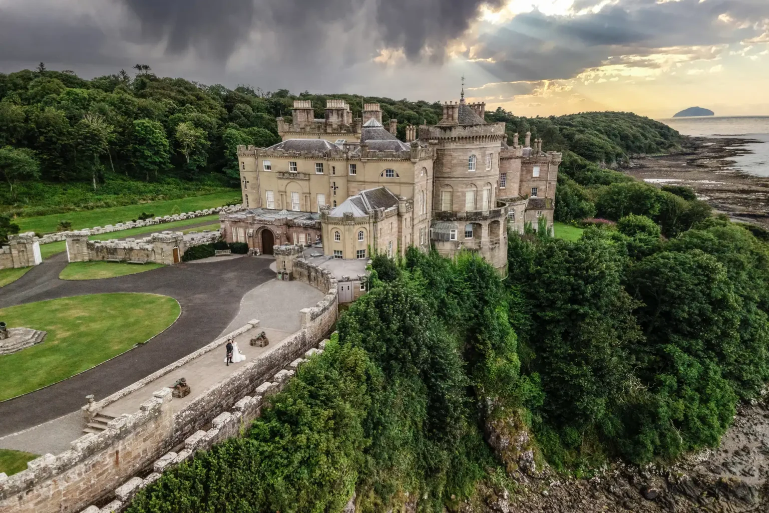 Aerial view of a bride and groom standing beside large, historic stone Culzean Castle with multiple turrets, surrounded by lush greenery, located near a rocky coastline. The sky is partly cloudy with sunlight breaking through.