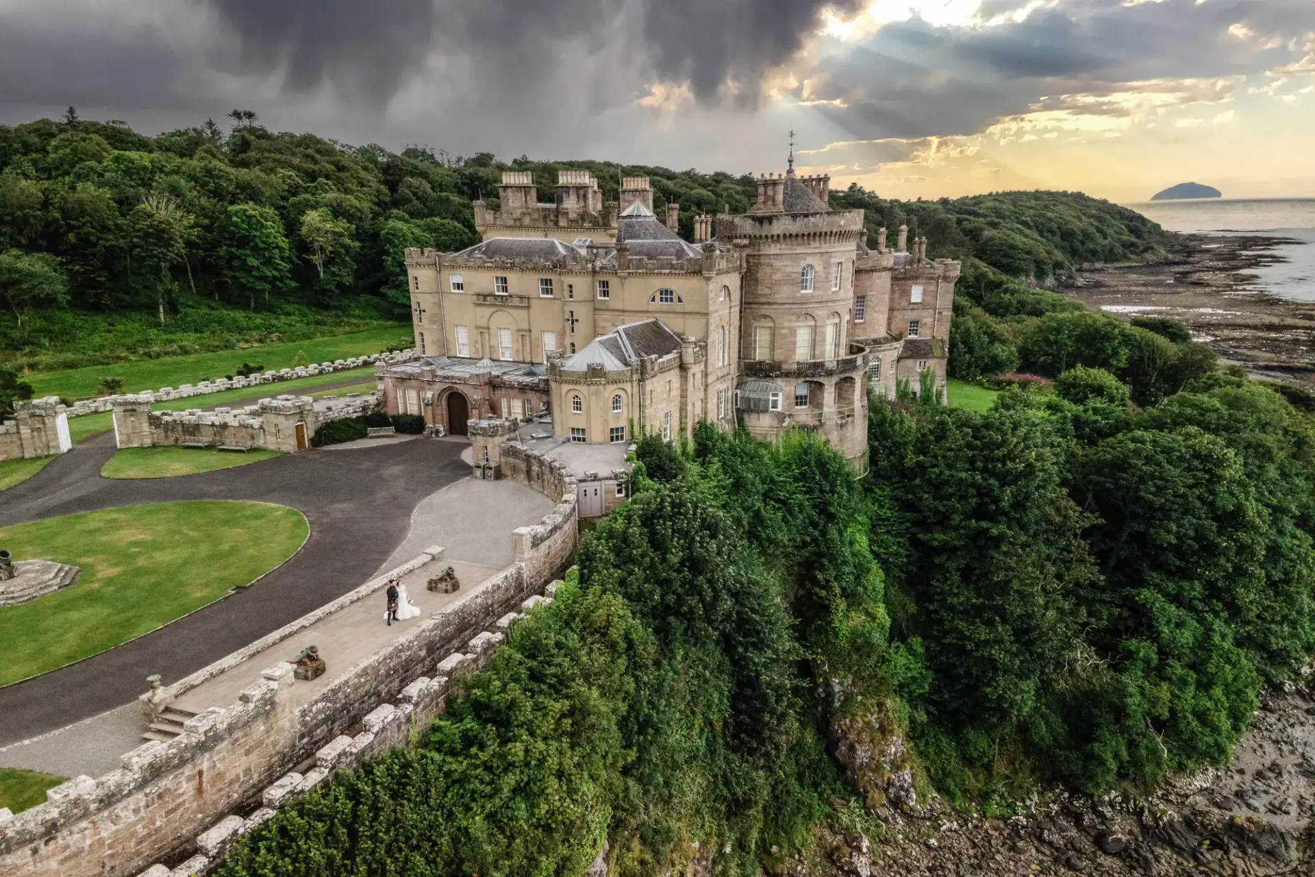 Aerial view of a bride and groom standing beside large, historic stone Culzean Castle with multiple turrets, surrounded by lush greenery, located near a rocky coastline. The sky is partly cloudy with sunlight breaking through.