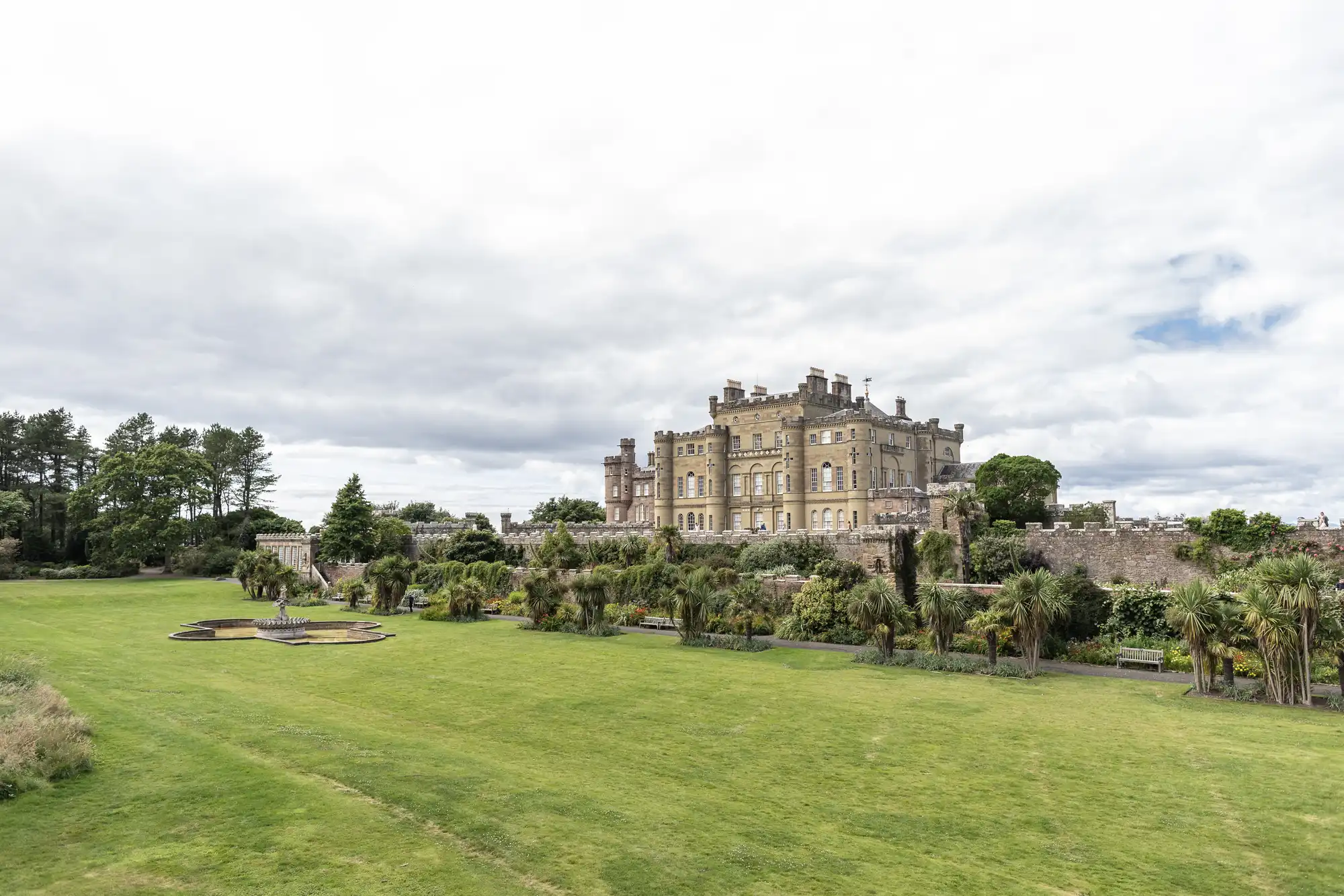 A large historic castle surrounded by green lawns and palm trees under a cloudy sky.