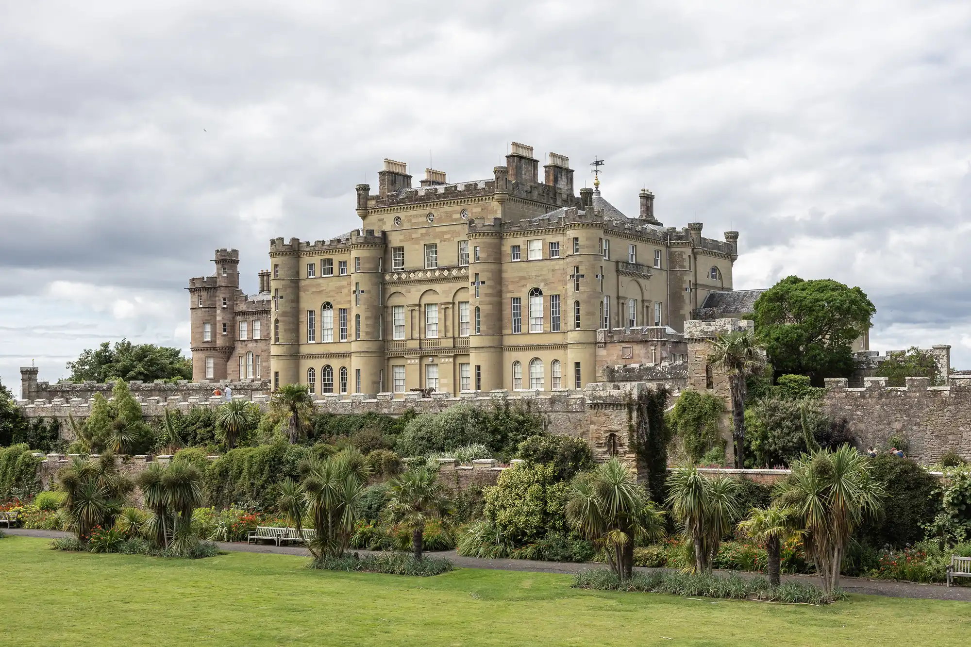 A large historic castle with a well-maintained garden in the foreground, surrounded by stone walls, against a cloudy sky backdrop.