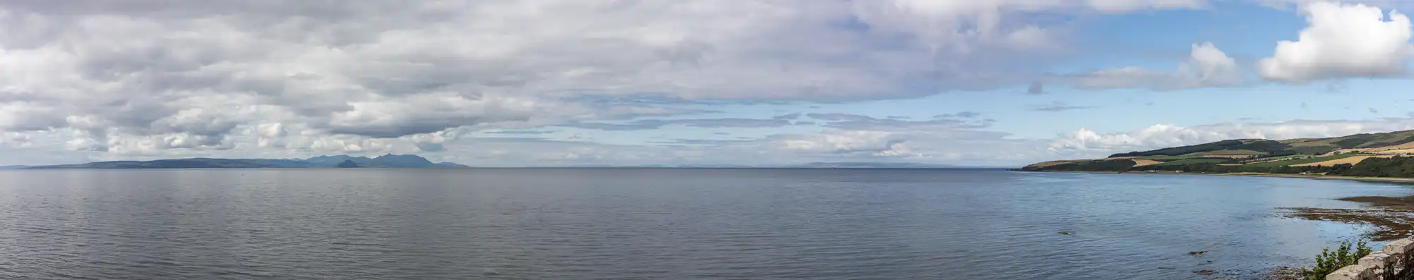 Panoramic view of a calm sea with a partly cloudy sky, distant mountains, and a coastline featuring greenery and hills on the right.