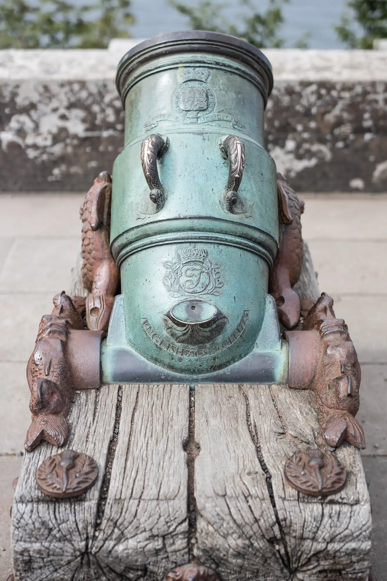 A bronze cannon with intricate carvings, mounted on a weathered wooden base, displayed outdoors near a stone wall.