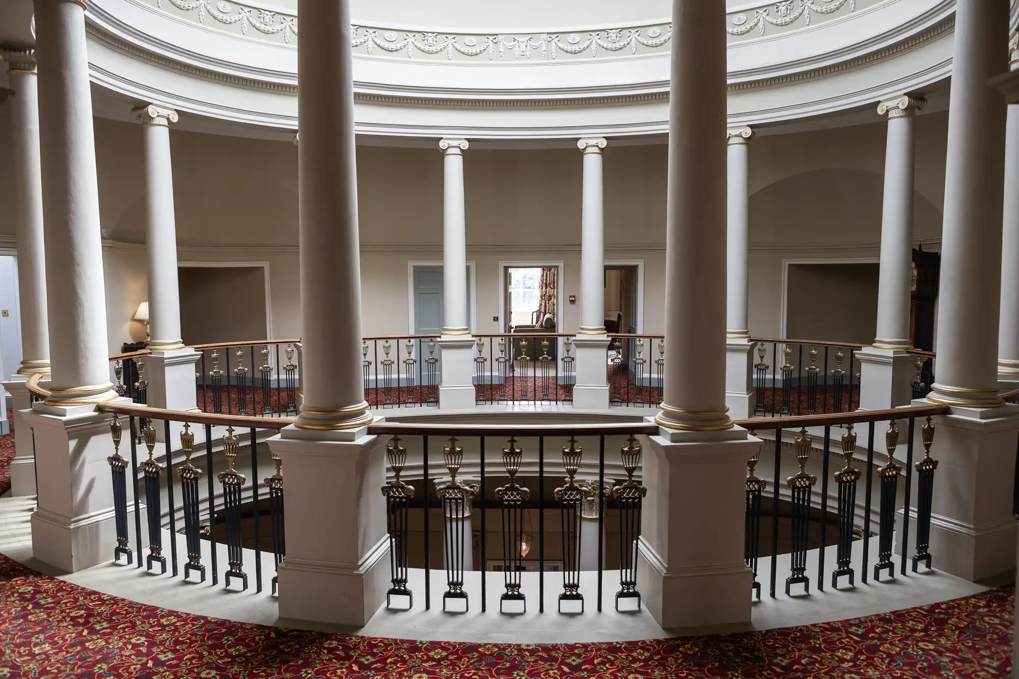 A circular balcony with columns and ornate railings surrounds an open space, framed by white walls and red carpeting with a patterned design.