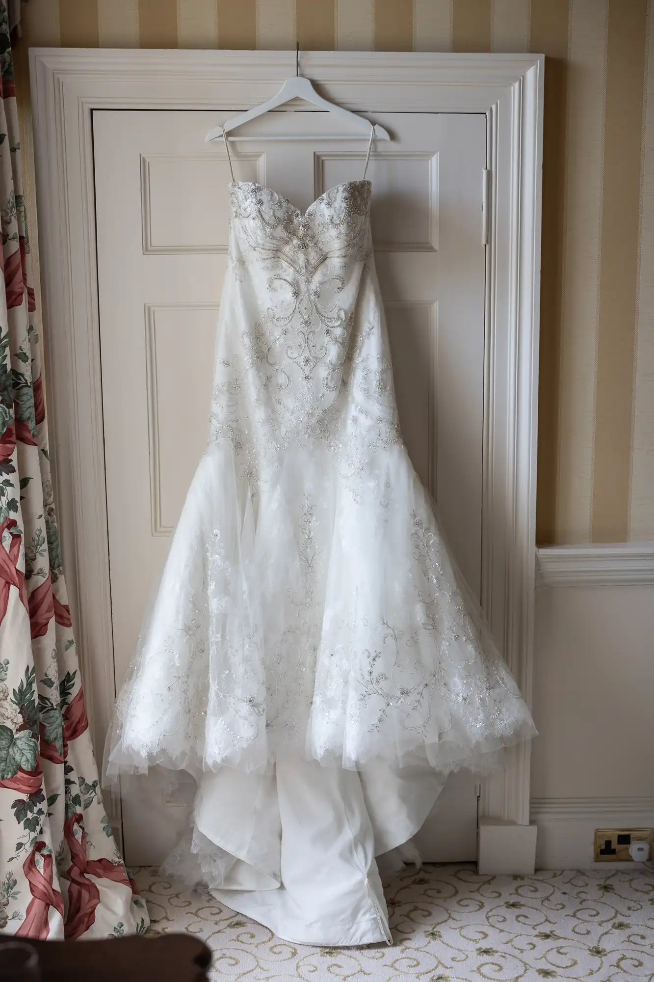 A white lace wedding dress with a fitted bodice and a flared skirt is hanging on a hanger on a door in a room with floral curtains and patterned wallpaper.