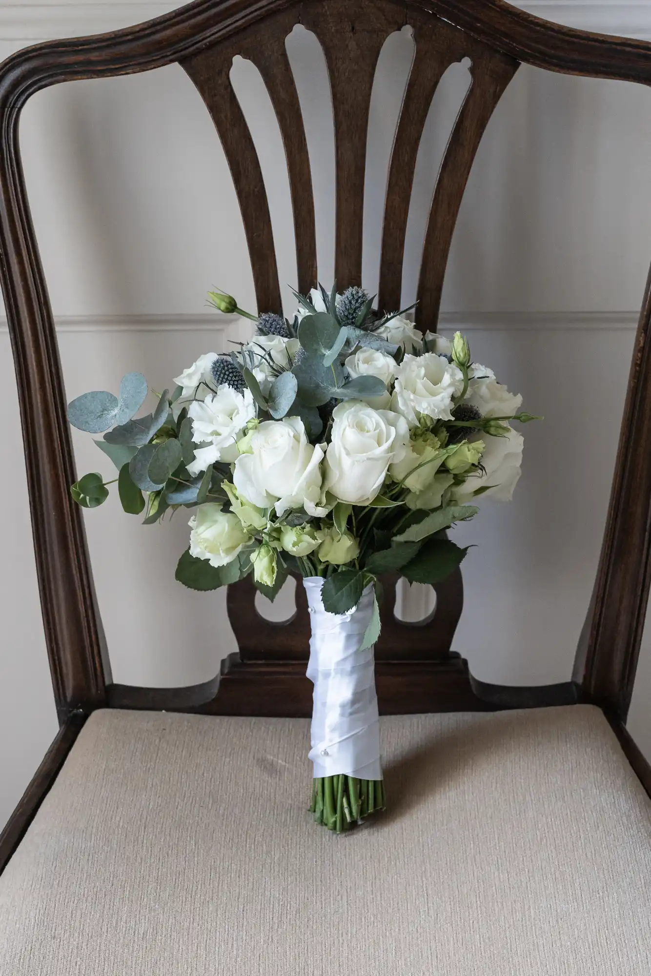 A wedding bouquet composed of white roses, greenery, and blue thistle, wrapped with white ribbon, placed on a beige cushioned wooden chair with a high back.