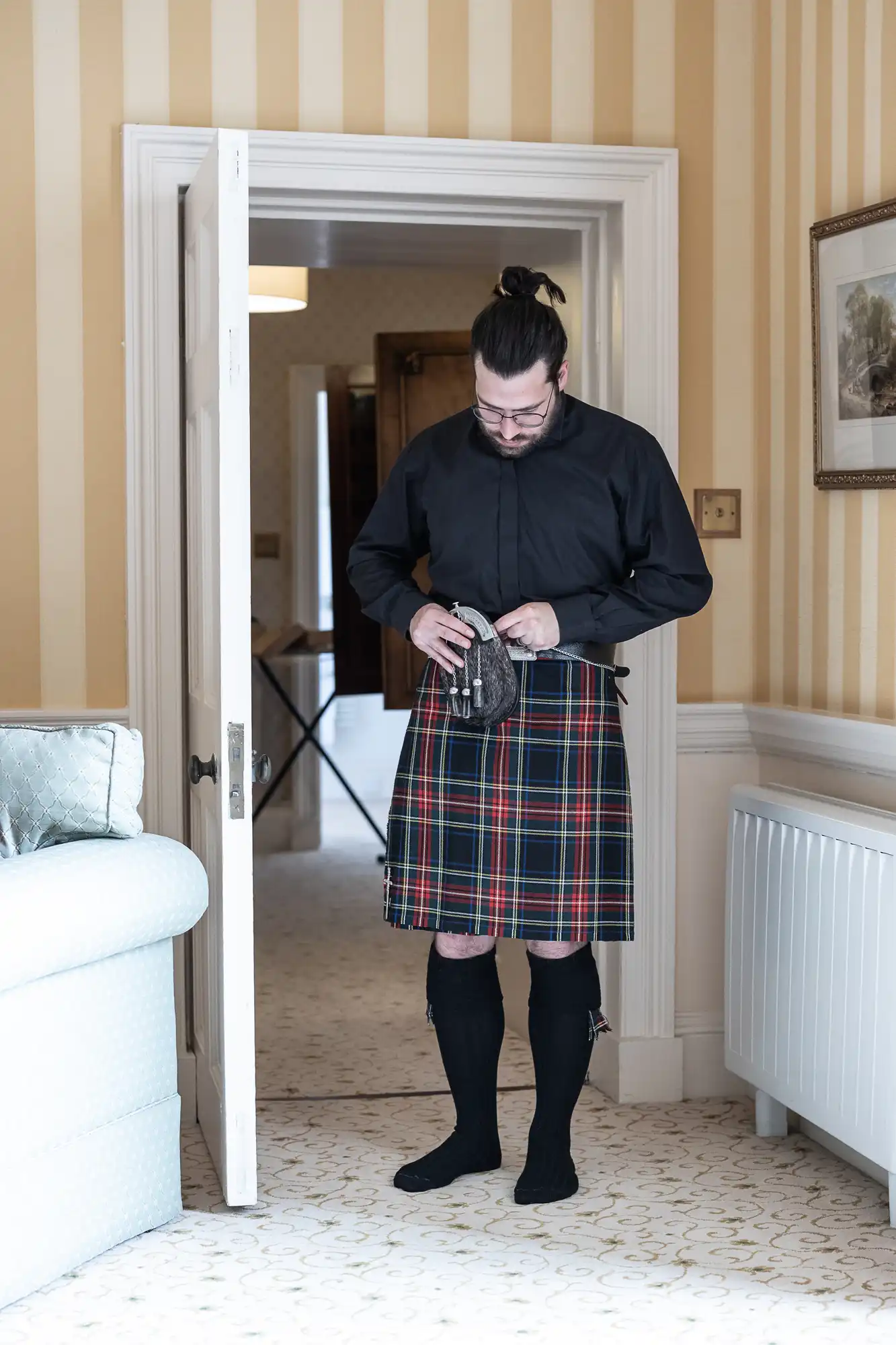 A man in a black shirt and traditional Scottish kilt stands in a room, adjusting his sporran. An ironing board and open door are visible in the background.