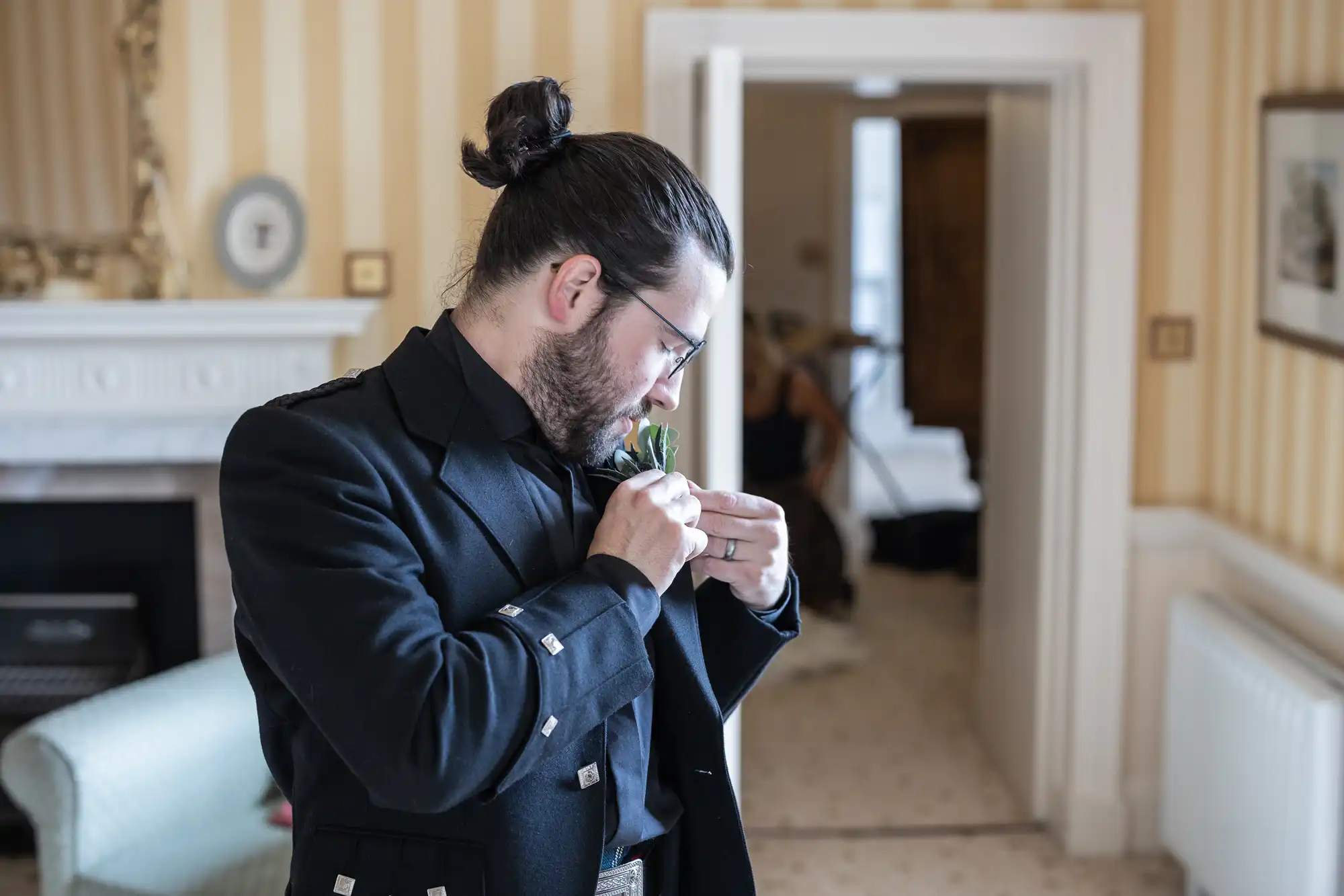 A man with a bun and glasses, wearing a black jacket, adjusts a boutonnière on his lapel in a room with striped wallpaper and an open door.