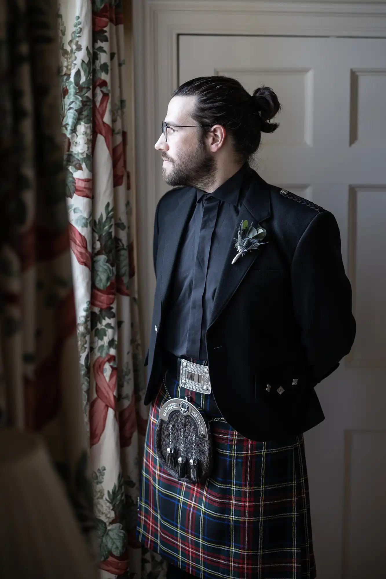 A man dressed in a traditional Scottish kilt outfit stands by a window, looking to the side. He is wearing a black jacket and shirt, with a sporran, tartan kilt, and brooch.