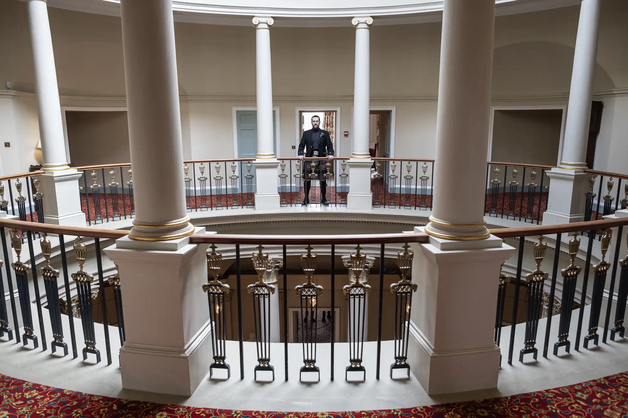 A person stands behind a black railing on the upper floor of a building with columns and a circular opening in the center. The lower floor is visible, and the carpeted area features a red and gold design.