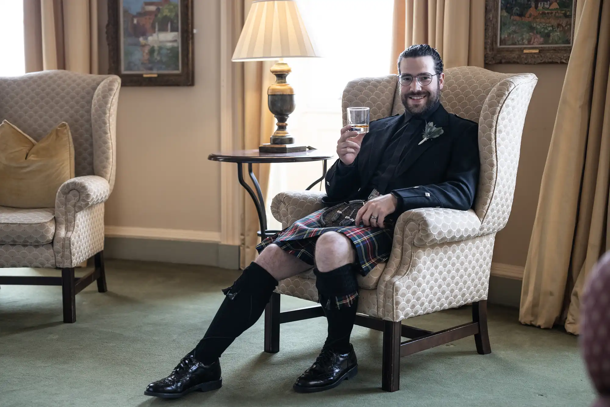 A man in a kilt sits in an armchair holding a drink, smiling. He is in a well-furnished room with a table and lamp beside him, flanked by another armchair and curtain.