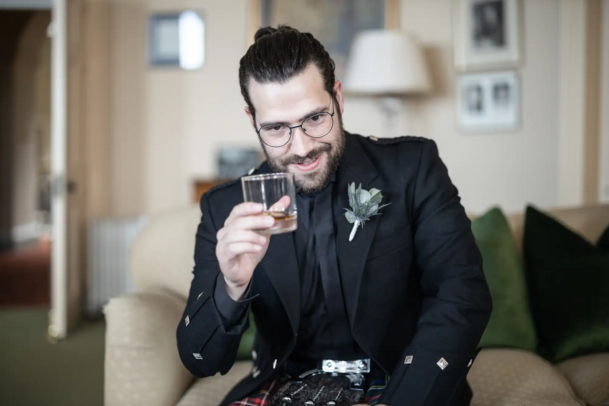 Man with glasses and beard wearing a kilt and suit jacket holding a glass of drink, sitting on a couch in a living room.