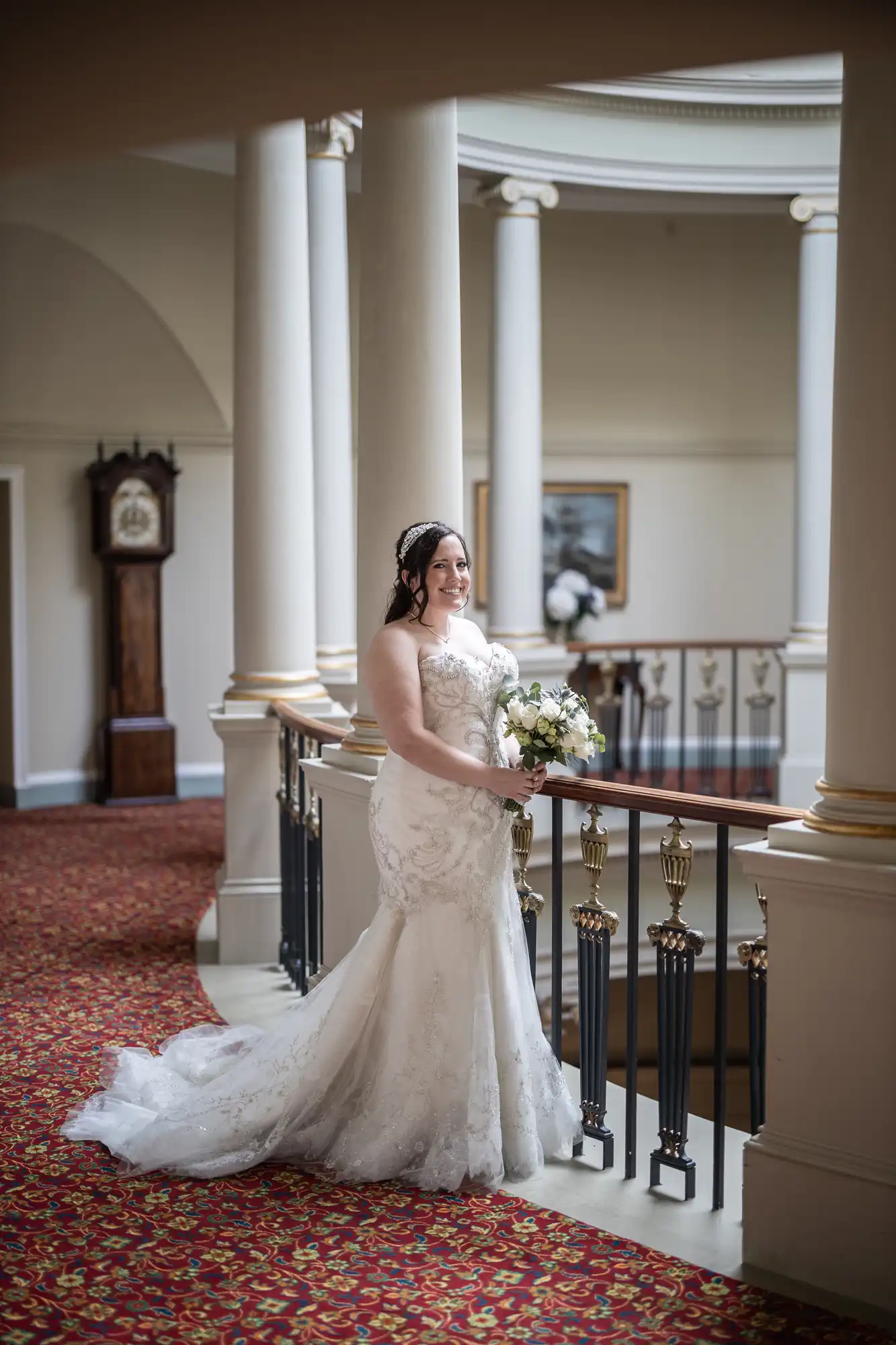 A bride in a white wedding dress stands by a balcony railing indoors, holding a bouquet of white flowers. The background includes columns, a clock, and a framed picture.