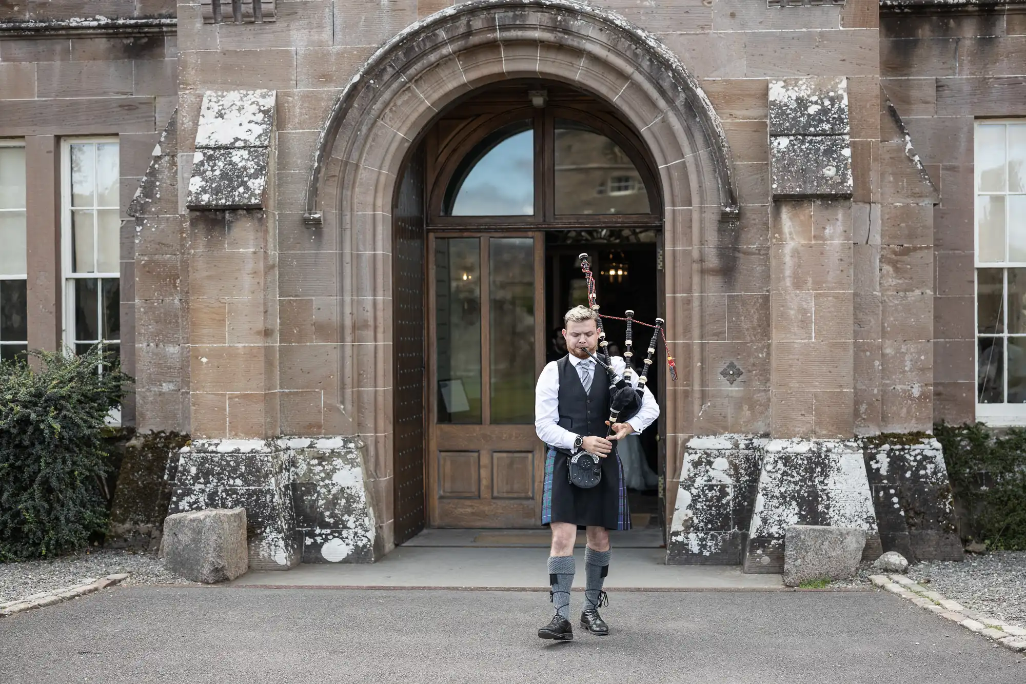 A man dressed in traditional Scottish attire is playing the bagpipes in front of an old stone building with an arched entrance.