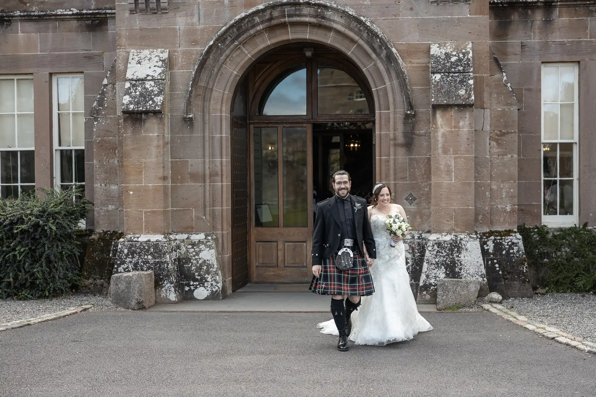 A man in a kilt and a woman in a white wedding dress smile as they walk arm-in-arm outside a large stone building with an arched doorway.