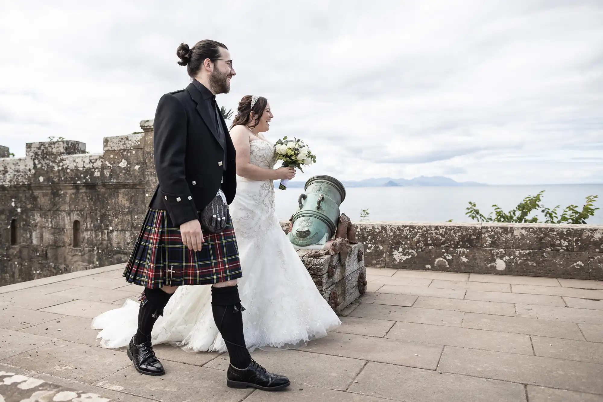 A bride in a white gown and a groom in a traditional Scottish kilt walk along an ancient stone structure overlooking the sea.