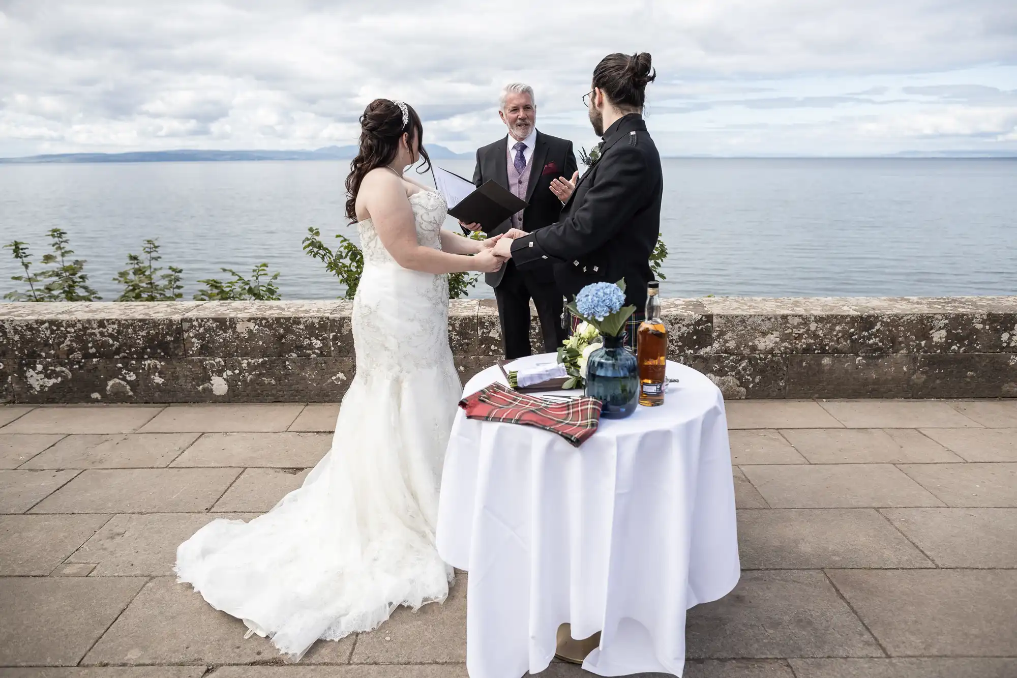 A couple stands before an officiant by the seaside, exchanging vows. They are dressed in wedding attire, with a table in the foreground holding flowers, a whisky bottle, and a tartan cloth.