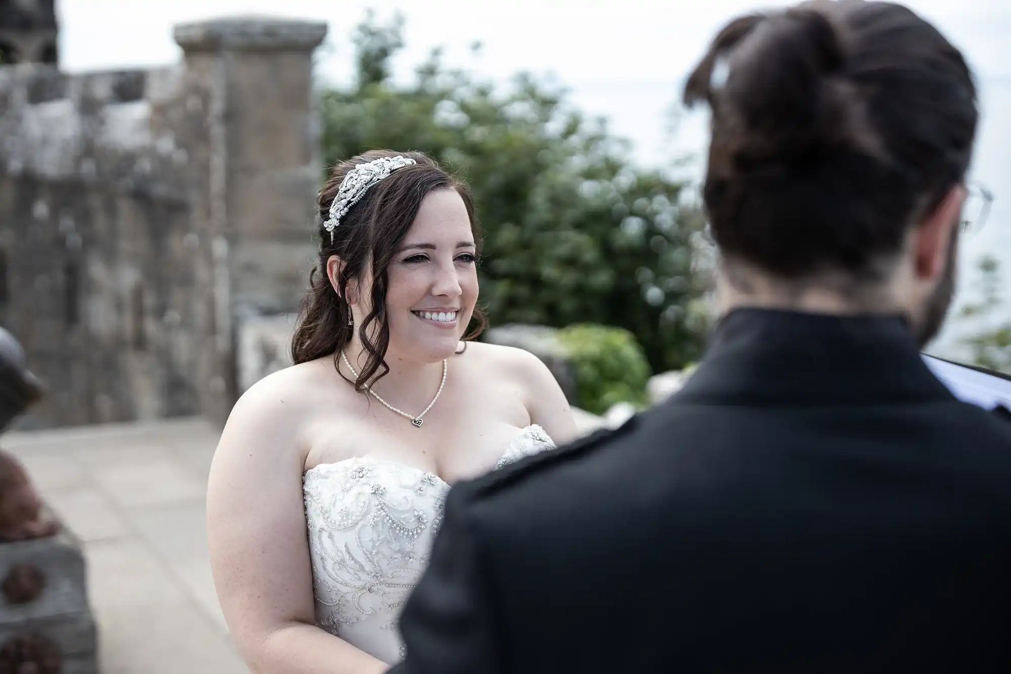 A bride in a strapless white wedding dress and tiara smiles, standing outdoors beside a stone structure. A person with dark hair, dressed in dark clothing, stands facing her.