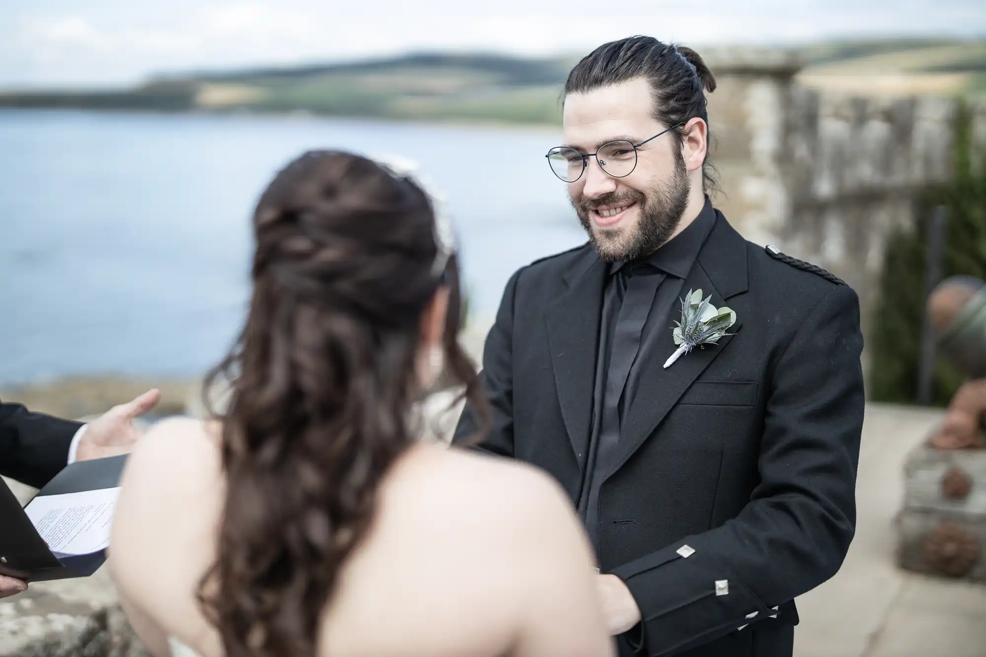 A man and woman stand facing each other during an outdoor ceremony near a body of water. The man, wearing glasses and a dark suit, smiles while holding the woman's hands.