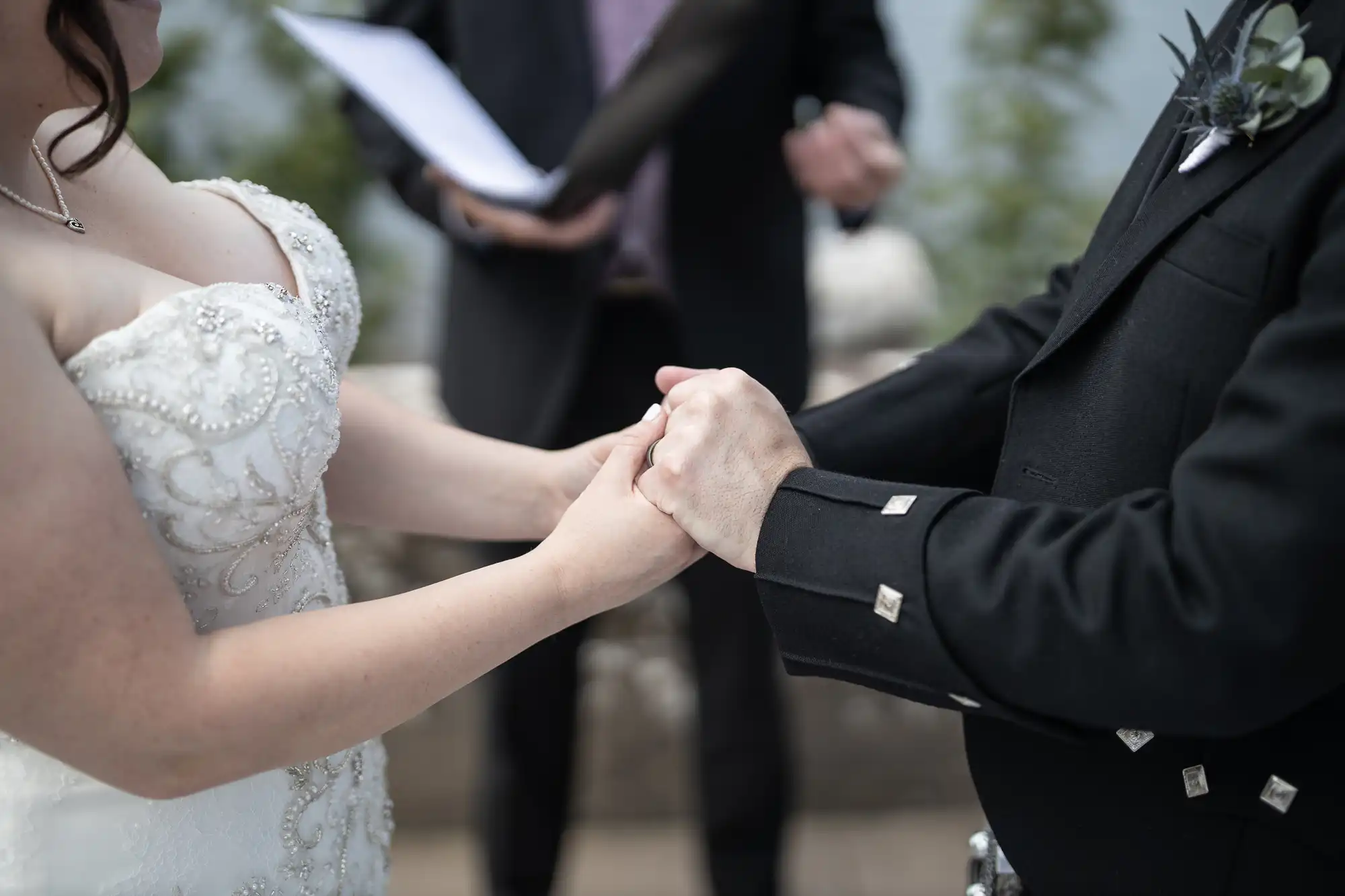 A couple is holding hands during a wedding ceremony. The bride is wearing a white dress with intricate beading, and the groom is in a black suit. An officiant stands in the background holding a document.