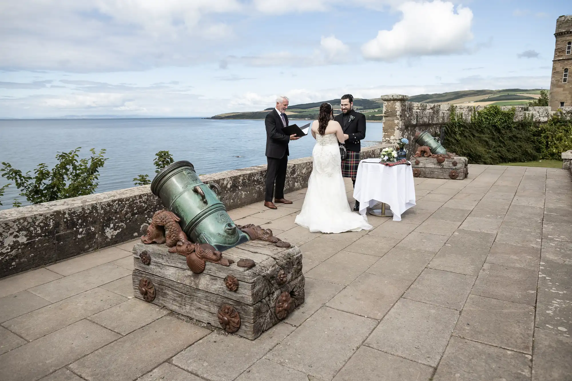 An outdoor wedding ceremony by the sea with three people; a couple getting married and an officiant. They stand near a table adorned with flowers and decorative items on a stone patio.